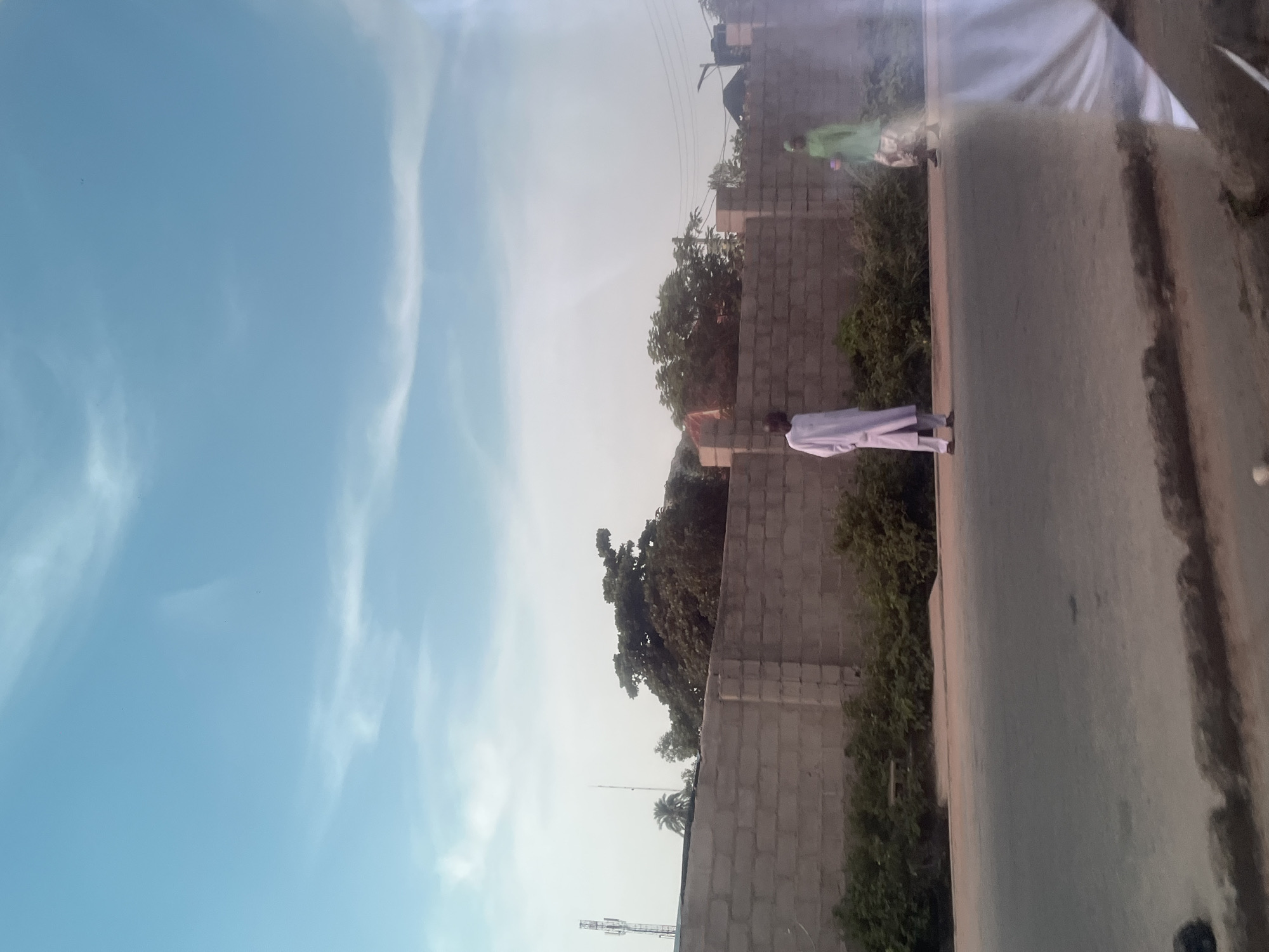 This media file depicts an outdoor scene with two individuals visible. The sky is a clear, bright blue with wispy clouds, suggesting it is daytime. The overall lighting indicates good weather. In the foreground, a person dressed in white clothing stands on a paved surface, possibly a road or walkway. Behind this person, a brick wall forms a barrier, with dense green foliage growing along its base. Further in the background, another individual is seen wearing a green garment, standing near the brick wall. The architecture of the brick wall, composed of cinder blocks, suggests a location in a developing area or a construction site. The presence of what appear to be communication towers in the distance provides additional context for the setting. The scene appears to be captured in Jalingo, Nigeria. There is no visible text in the image.