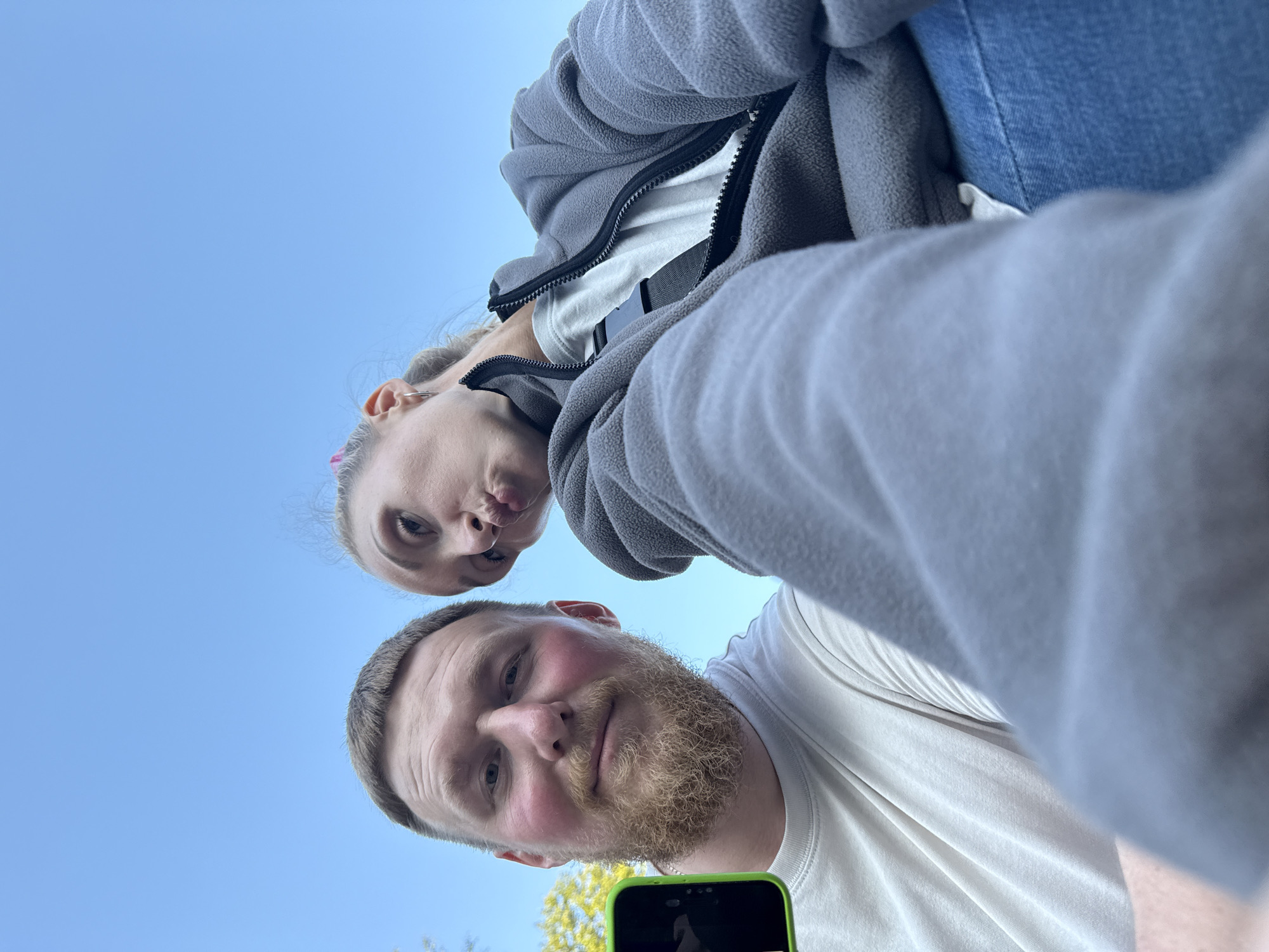 A low-angle, selfie-style shot shows two individuals outdoors against a clear blue sky. In the foreground, a man with a beard and a white t-shirt is holding a smartphone with a green case, suggesting he is taking the photo. To his right, a woman with blonde hair pulled back and a pink accessory is leaning in, making a pouty expression. She is wearing a grey zip-up jacket over a white t-shirt and blue jeans. The background is dominated by the bright sky, with hints of green foliage visible on the left side. The setting appears to be a park or outdoor recreational area in the city of Perm, Russia.