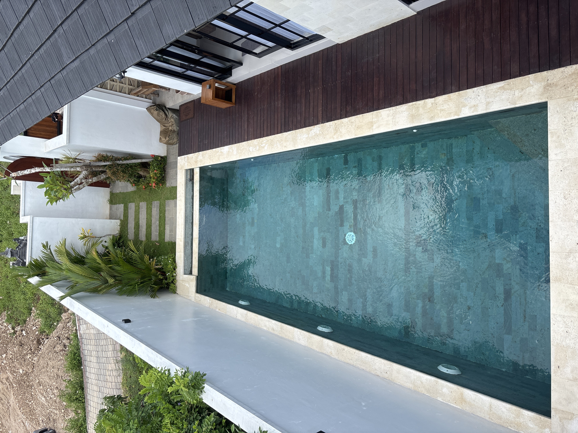 This high-angle photograph depicts a swimming pool and surrounding architecture in Ungasan, Indonesia. The pool, filled with turquoise water and featuring a tile bottom, is bordered by light-colored stone or tile. Dark brown wooden decking extends from the pool's edge towards a building with a dark grey tile roof and black-framed windows. A white, modern-style building is situated adjacent to the pool, surrounded by green landscaping, including palm trees and a lawn with evenly spaced stone pavers. The surrounding environment includes areas of dirt and other vegetation, indicative of an outdoor setting.