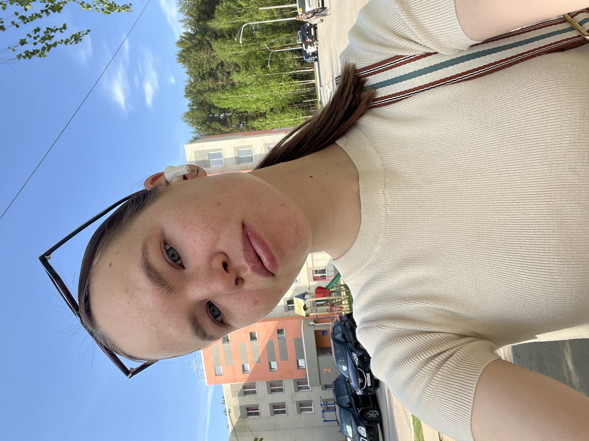 The image shows a woman taking a selfie outdoors. She has light skin, brown hair pulled back, and is wearing a light-colored top with a striped strap over her shoulder. Sunglasses are perched on her head. She appears to be on a street in Chaikovsky, Russia, based on the presence of multi-story buildings and vehicles in the background. Trees and blue sky with some clouds are also visible.
