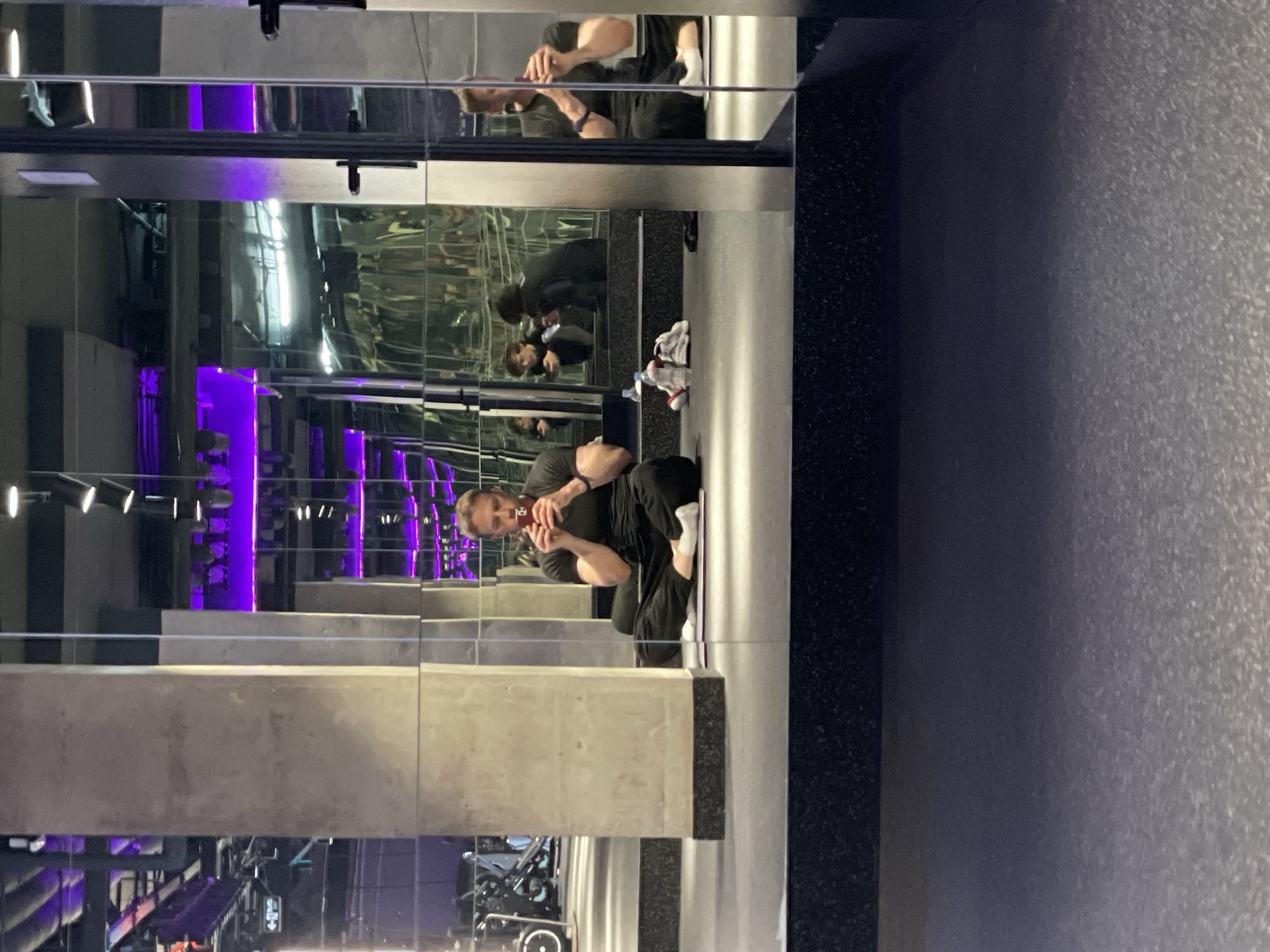 A man is reflected in a mirrored wall in what appears to be a gym or fitness studio. The man is wearing dark clothing and is holding a phone in his hands, seemingly taking a photo. The reflection shows multiple repetitions of the man and the surroundings. In the background, there are gym equipment, lights, and purple neon lighting, suggesting a modern or upscale fitness facility. The location is hinted at as Murino, Russia, due to the mention of the city in the prompt.