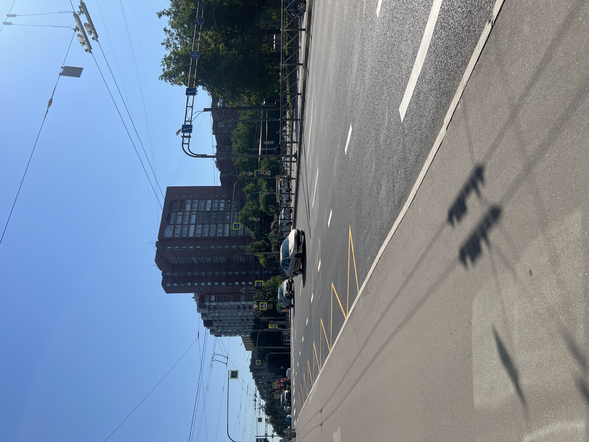 The image presents an urban street scene in Saint Petersburg, Russia, captured on a clear, sunny day. The perspective is along a multi-lane asphalt road, stretching from the foreground into the distance. The road features white dashed lane markings, a solid white line, and distinctive yellow zigzag markings, possibly indicating a bus stop or a no-parking zone, on its right side. Long, distinct shadows, cast by unseen objects, stretch across the road surface, suggesting either morning or late afternoon.
To the left of the roadway, a metal barrier or fence separates it from a pedestrian area or green space. Behind this barrier, a line of lush green trees partially conceals multi-story residential or commercial buildings, characterized by numerous windows and dark brick or stone facades.
Overhead, a dense network of wires, characteristic of a trolleybus or tram system, traverses the clear blue sky, supported by numerous utility poles and structures. Traffic light gantries and various road signs are also visible. Among the signs, several yellow square signs with black pedestrian crossing symbols are prominent along the left side of the street, alongside blue rectangular signs with white directional arrows.
A few vehicles are present on the road. A light-colored car is visible in the mid-ground, with another darker vehicle behind it. Other cars are parked or moving slowly further down the street on the left. The scene depicts a typical, busy urban environment, with no specific event unfolding beyond routine street activity. No discernible text is visible on any signs or buildings beyond the symbolic representations on traffic signs.