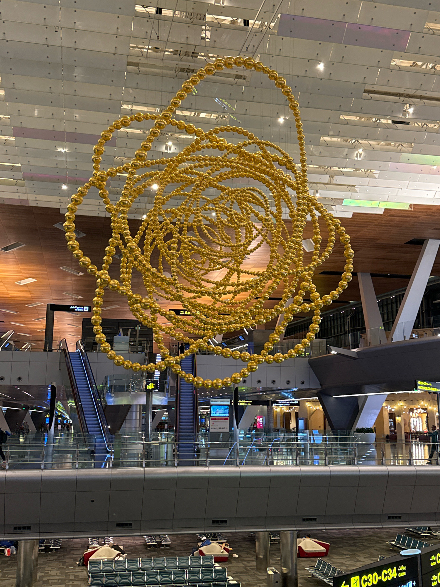 This image shows the interior of Hamad International Airport in Doha, Qatar. A large, intricate golden sculpture made of spherical beads hangs from the ceiling in the center of the frame. The sculpture is spherical and appears to be made of interconnected rings. Below the sculpture, there are multiple levels of the airport visible, with escalators, walkways with glass railings, and seating areas. People are visible walking on the upper levels and resting in the seating areas on the lower level. The environment appears to be a modern, spacious airport terminal with a high ceiling and wooden paneling. Signs in both English and Arabic are visible throughout the terminal, indicating gates and directories. The lighting is a mix of natural light filtering through the ceiling panels and artificial lights.