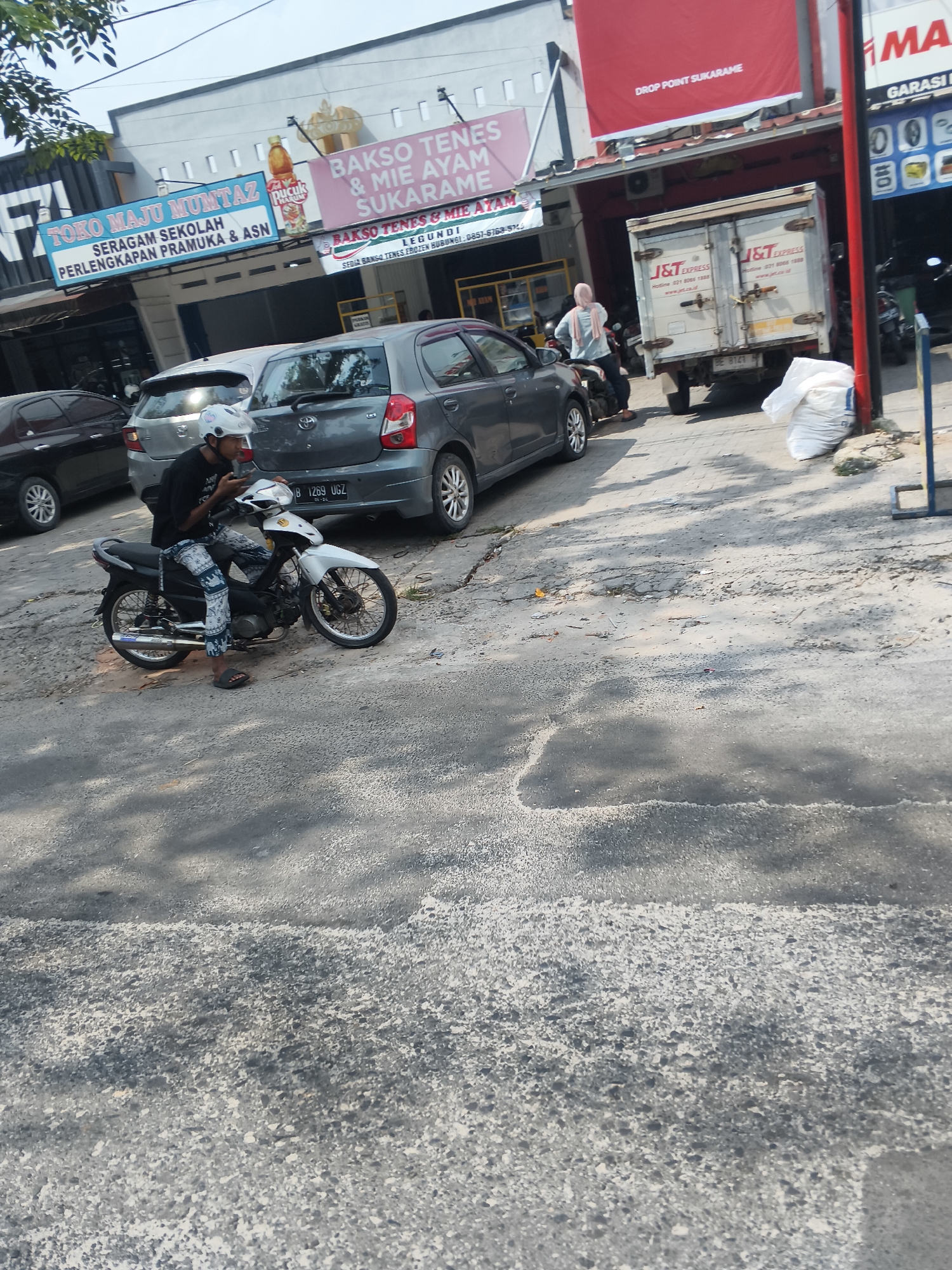 The image depicts a street scene in Bandar Lampung, Indonesia, likely a commercial area. In the foreground, a person is seated on a white motorcycle, holding a mobile phone. The motorcycle is parked on a cracked asphalt road. Behind the motorcycle, a gray car is visible. Further back, a white J&T Express delivery truck is parked. Several buildings line the street, displaying various signs. One sign reads "TOKO MAJU MUMTAZ SERAGAM SEKOLAH PERLENGKAPAN PRAMUKA & ASN." Another sign, in pink, advertises "BAKSO TENES & MIE AYAM SUKARAME." A red banner above the J&T truck indicates "DROP POINT SUKARAME." The environment appears to be outdoors, with daylight casting shadows on the road. The overall scene suggests a typical day in a busy Indonesian city.