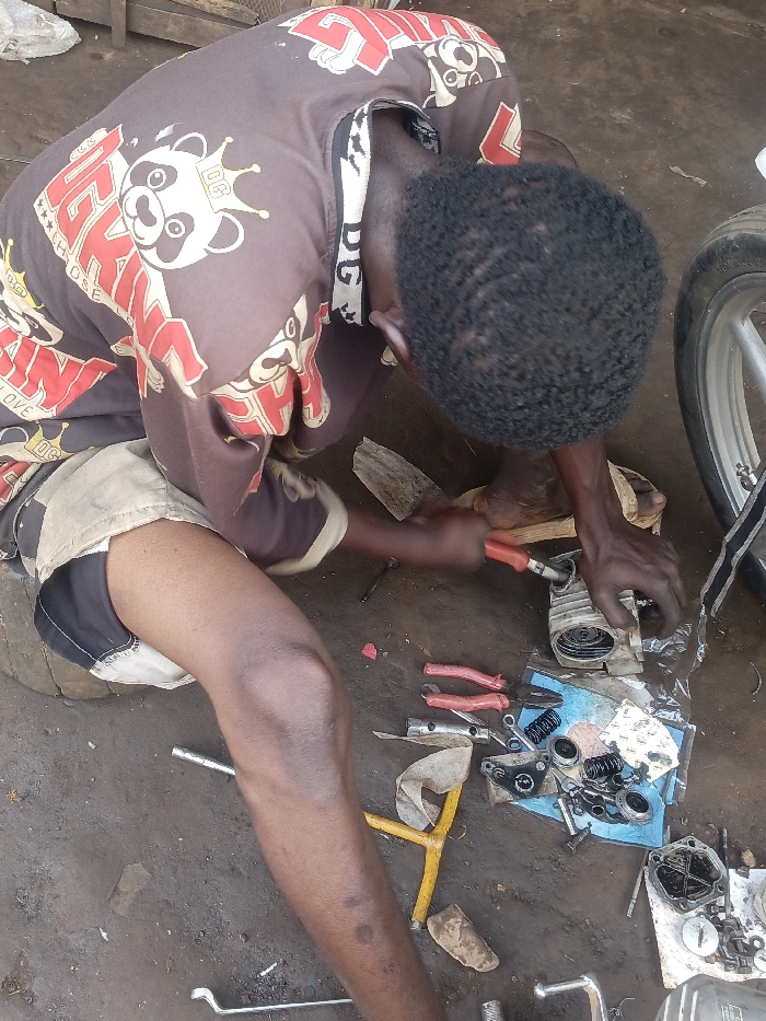 A person crouches on a dirt floor, working on what appears to be a motorcycle engine part. He uses a red-handled tool to manipulate the object. Several other tools, including pliers and wrenches, are scattered around along with disassembled engine components. The person wears a brown patterned shirt, khaki shorts, and sandals. A motorcycle wheel is visible on the right side of the frame.