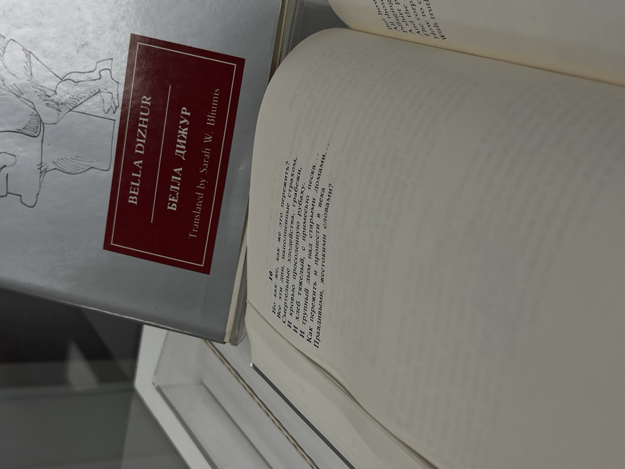 This close-up shot, taken in Yekaterinburg, Russia, features two books. In the foreground, an open book displays text on both pages. The left page shows Russian Cyrillic script, while the right page presents its English translation. The visible Russian text is numbered "10" and begins with the lines: "Но как же, как же это пережить? / Все эти дни, наполненные страхом, / Смертельные злодейства, грабежи, / И кровью просоленную рубаху. / И хлеб тяжелый, с примесью песка... / И трупный дым над старыми домами. / Как пережить и пронести в века / Правдивыми, жестокими словами?" The partial English translation visible on the right includes phrases like "But how," "All those," "Pillaging," "A shirt p," "And the," "The corp," "How to c," and "With truth."
Beneath the open book, the cover of another book is visible, partially obscured. This book, resting face-up and slightly tilted, has a light gray cover adorned with a faint line drawing of a figure. A rectangular, reddish-brown label is affixed to its cover, bearing the titles "BELLA DIZHUR" in English and "БЕЛЛА ДИЖУР" in Russian, followed by "Translated by Sarah W. Bliumis." The books appear to be on a flat, light-colored surface, possibly a shelf or table, within an indoor setting. The lighting is soft and even, suggesting ambient indoor illumination. No people are visible, and the overall atmosphere is quiet and academic, focusing on the written word and translation.
