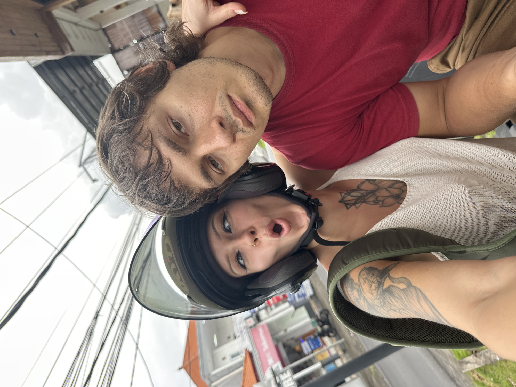 A selfie of a man and a woman on a motorcycle in Munggu, Indonesia. The man, in a red shirt, is behind the woman who is wearing a helmet. The background shows buildings, power lines, and vegetation. The photo is angled. The woman is wearing a light colored blouse and an olive-green jacket. She has tattoos on her arm and upper chest. The man has brown hair.
