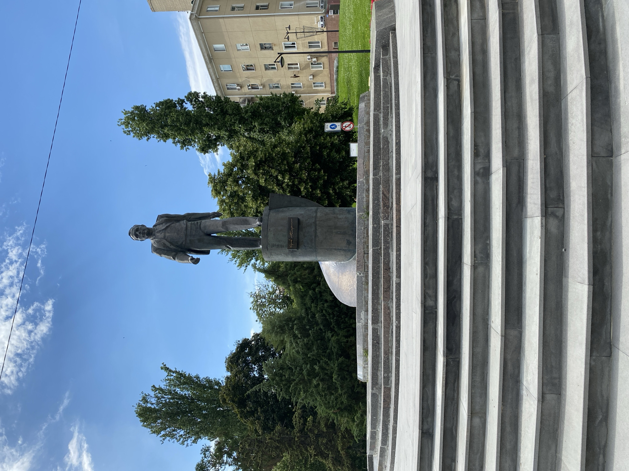 A bronze statue of a man in a suit stands on a pedestal in an outdoor plaza on a sunny day. The statue is the central focus of the image, positioned on a raised platform with stairs leading up to it. Behind the statue, lush green trees form a dense backdrop, with a light-colored apartment building visible to the right. The sky is blue with scattered white clouds. The plaza appears to be made of stone, with the stairs and platform constructed from light-colored granite. A few signs and a lamppost are present on the right side of the image. The overall scene suggests a public space with a monument.