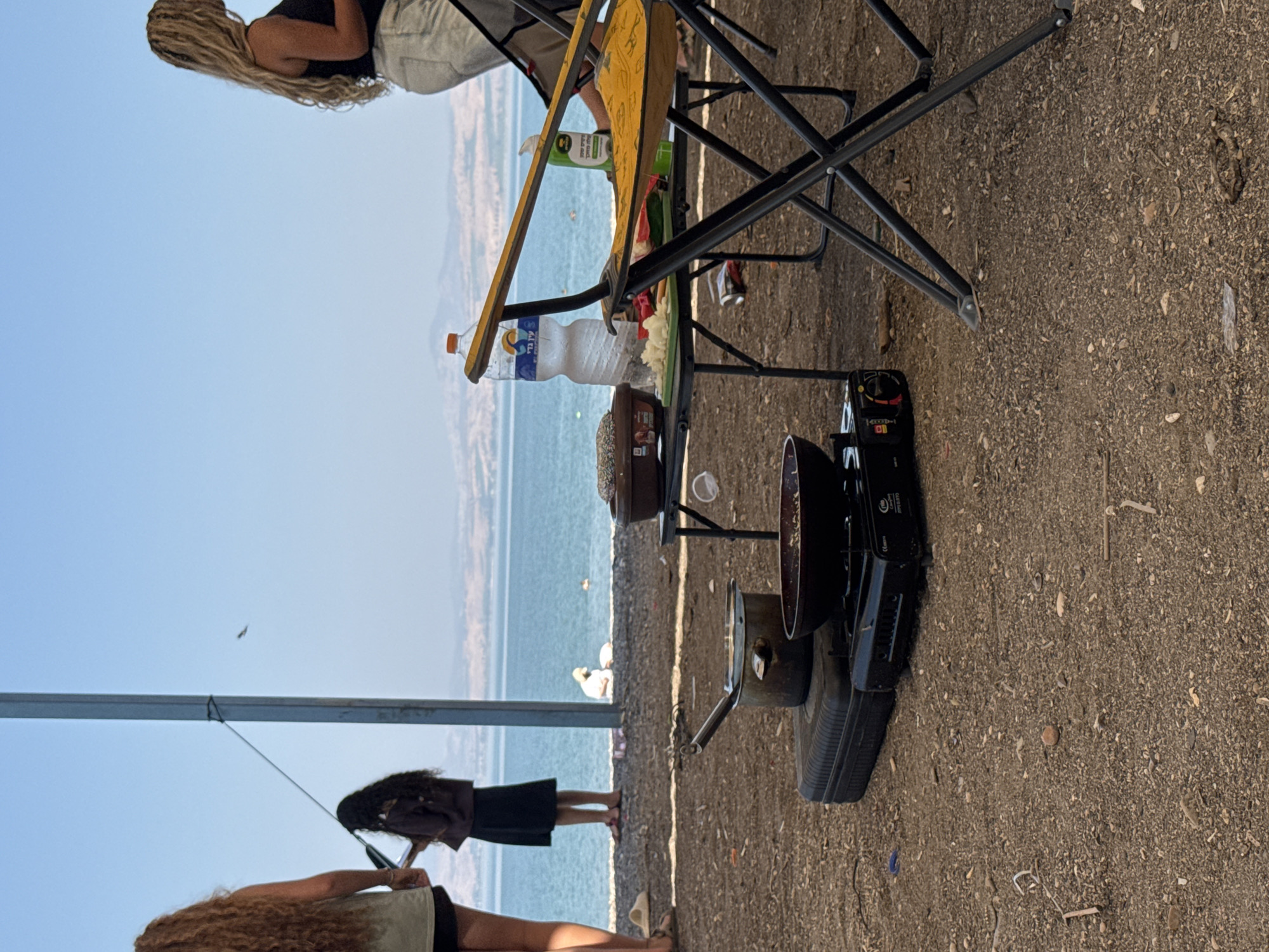 This image depicts an outdoor scene at a campsite by the Sea of Galilee in the Golan Regional Council, Israel. In the foreground, a portable camping stove is set up on a black case, with a pot and a frying pan on top. A black metal folding chair with a yellow seat and back is positioned to the right, partially obscuring a table. On the table, there is a cutting board with chopped onions and other ingredients, a bottle of water, and a green bottle, likely a cleaning product. In the middle ground, two women are standing near a large metal pole, and a third woman is visible further in the background, near the water's edge. People are also seen swimming in the Sea of Galilee. The background consists of the calm blue water of the lake and a hazy shoreline with hills beyond. The ground is covered with dirt, small stones, and scattered debris.