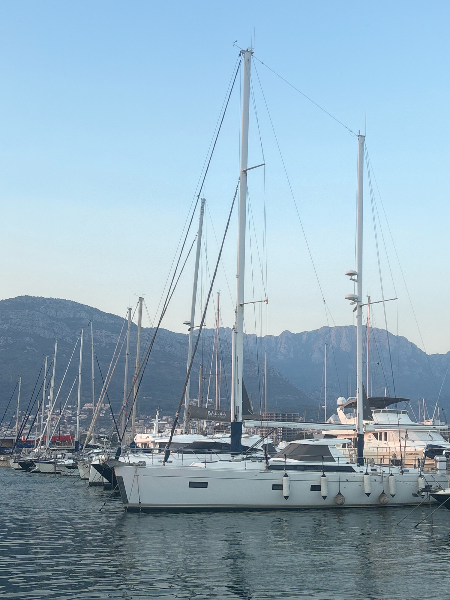In Bar, Montenegro, this image captures a marina filled with sailboats and yachts docked on calm, rippling water. The sky is a clear, light blue, fading into a hazy tone near the horizon. In the background, a range of rugged, rocky mountains forms a natural backdrop to the scene. Several sailboats, with their tall masts and rigging, are prominently featured in the foreground and mid-ground, some bearing the names "BALI44" and "NAMASTE." Other vessels, including a larger white yacht, are also visible. The overall impression is one of a peaceful harbor bathed in soft light, likely during the early morning or late afternoon. There are no visible people in the image.