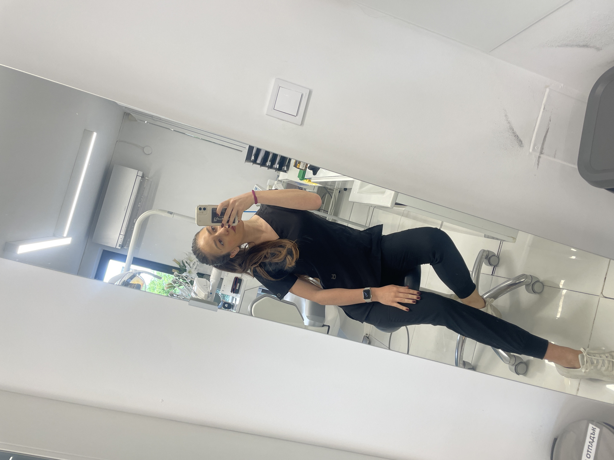 A woman is taking a selfie in a mirror. The location is likely a dentist's office, as dental equipment is visible. The woman is wearing a black top and black pants, and she's seated on a rolling chair. She is holding a phone up to take the picture. The reflection shows other dental equipment, an air conditioner, a light fixture, and a waste bin with writing in Bulgarian. The reflection is in a clean, modern space with white walls and tile flooring. Based on the language on the waste bin, the city is likely Sofia, Bulgaria.