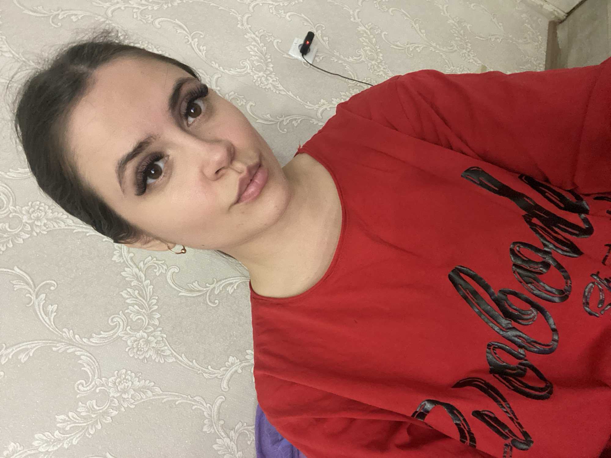 A woman is shown in a medium close-up shot, angled to the left. She has dark hair pulled back, with a fair complexion and wearing heavy false eyelashes. She is wearing a red t-shirt with black cursive lettering on it. The background is a light-colored wall with a floral pattern. A power outlet with a charger plugged into it is also visible on the wall. The location of the image is unknown, the reference to Chaikovsky, Russia, is not relevant to the image.