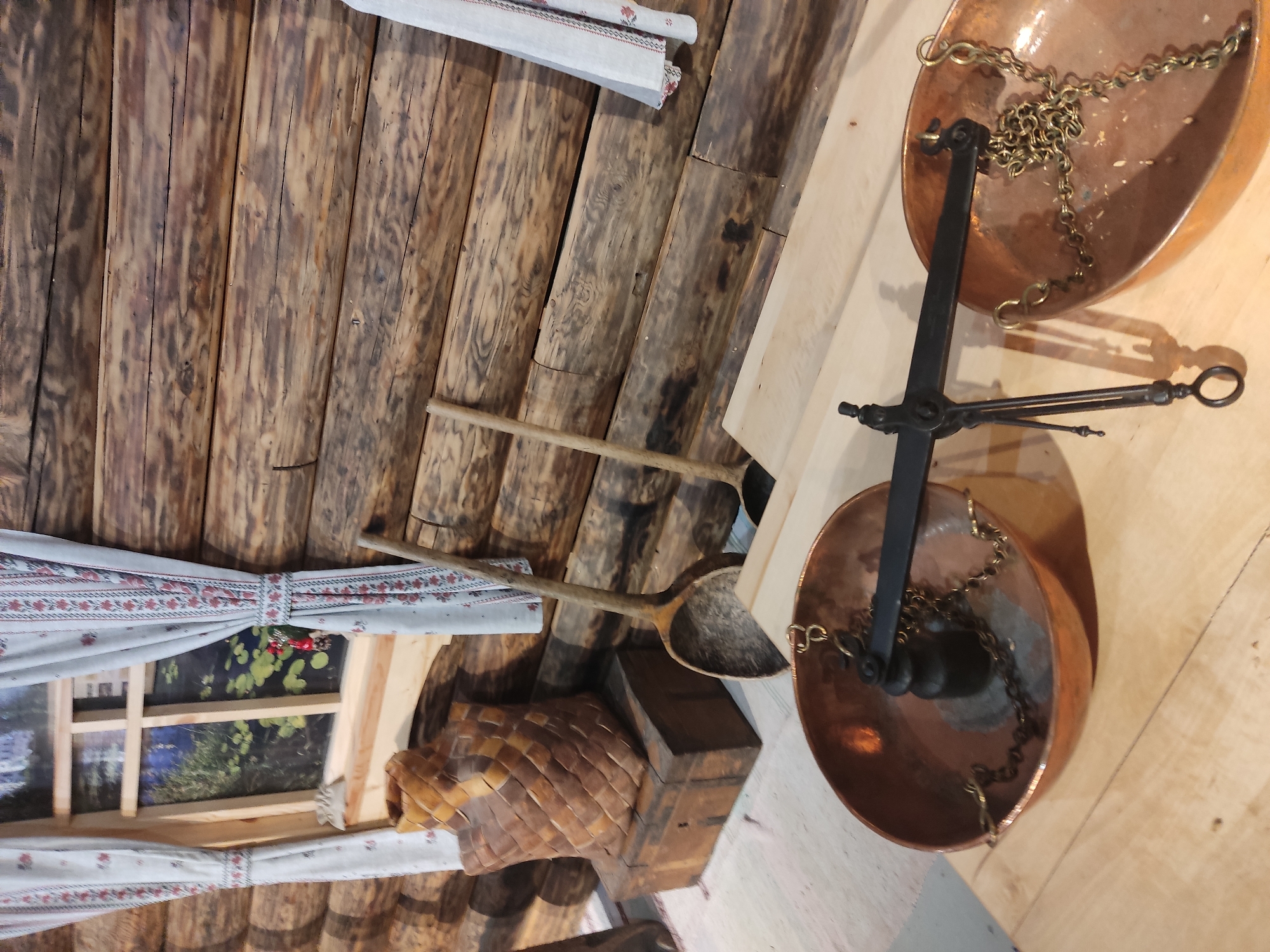 The image shows a rustic interior, possibly a museum exhibit, in Pskov, Russia. The walls are constructed of rough-hewn logs. A window is partially visible, covered by a simple curtain with a floral design. In the lower left, there is a birch bark container or basket. Two wooden spoons or ladles rest against the wall, near the basket. A set of antique copper scales is positioned prominently on the right side of the image. The scales have two copper pans suspended by chains and a central balance mechanism.