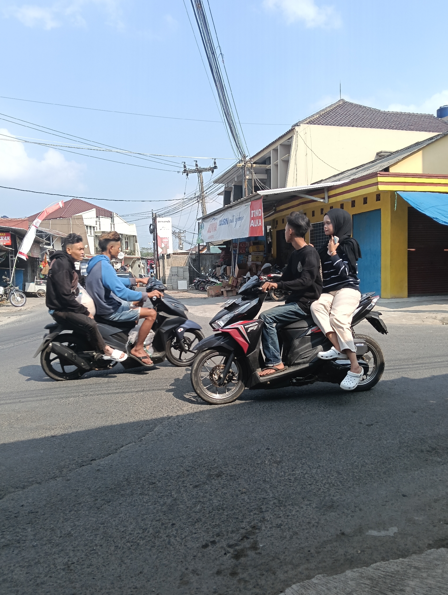 In Puspahiang, Indonesia, on a clear day, two motorcycles are seen on a paved road. The motorcycle on the left carries two young men. The driver, wearing a black hoodie and dark pants, has a white plastic bag on his lap. The passenger, in a blue hoodie and denim shorts, wears white sandals. They are moving to the left.
The motorcycle on the right is ridden by a young man in a black long-sleeved shirt and blue jeans. Behind him, a young woman wearing a black hijab and a striped long-sleeved shirt, along with light-colored pants, is seated. She wears white Crocs. This motorcycle is also moving to the left.
The environment includes buildings with yellow and brown facades, some with awnings and closed shutters. Electrical wires are strung across the sky, supported by utility poles. The road surface shows some cracks and discoloration. In the background, parked motorcycles and stacked building materials are visible near the shops.