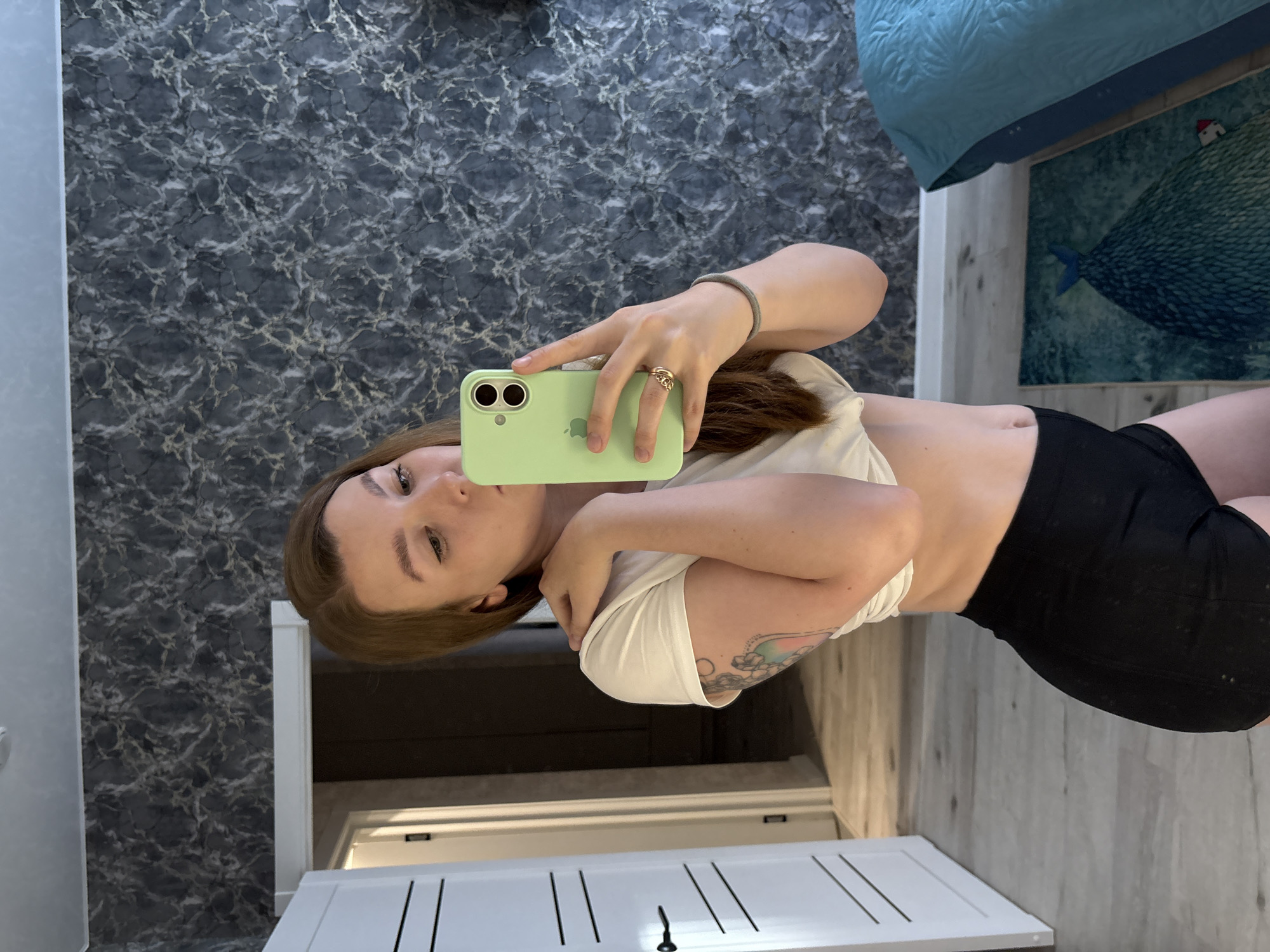 A young woman with long brown hair and a white t-shirt tied at her waist is taking a mirror selfie. She is wearing black shorts and has a tattoo on her left arm. She is holding a light green smartphone in her right hand, and a ring is visible on her right ring finger. She is standing in a room with dark gray, marble-patterned wallpaper. To her left, there is a white door with black horizontal lines and a black handle. In the background, a blue bed with a textured comforter is visible. The floor is light-colored wood. The image was taken in a bedroom in Izhevsk, Russia.