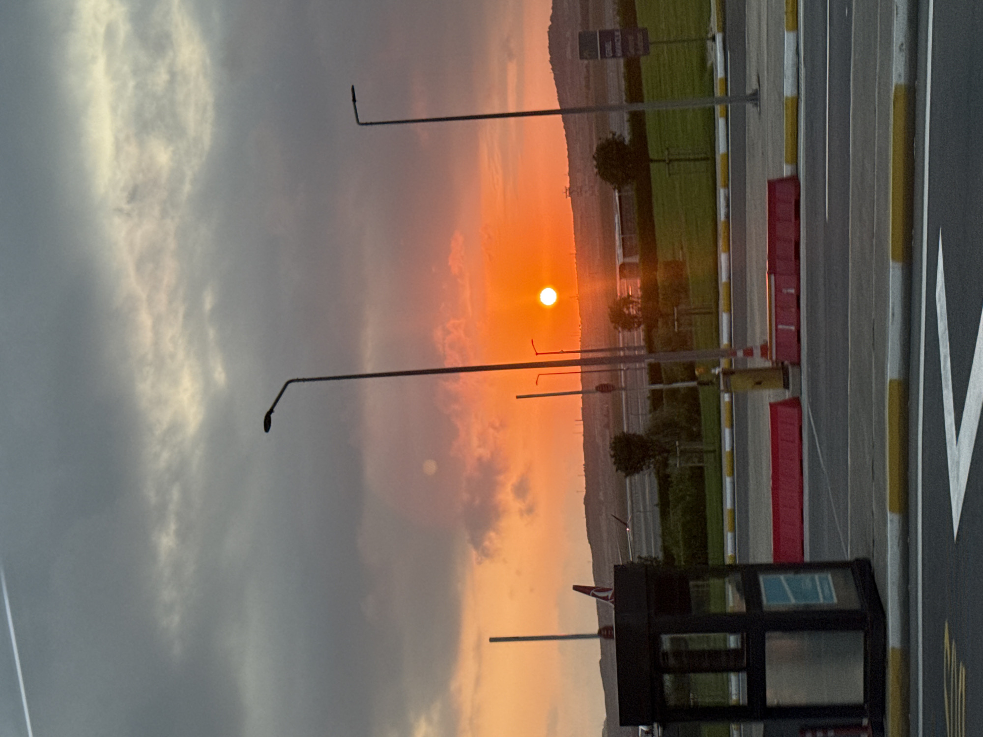 This is a photograph taken at Arnavutköy, Turkey, during sunset. The sky is filled with dramatic clouds, ranging in color from dark gray to vibrant orange and pink hues, indicative of the setting sun. The sun itself is a bright, luminous orb low on the horizon, casting a warm glow across the scene.
In the foreground, there is a road with clear lane markings and a yellow and white striped curb. A dark, box-like structure, possibly a security or toll booth, is positioned on the left side of the frame. Red traffic barriers are visible behind it. Several lampposts are strategically placed throughout the scene, some illuminated by the fading daylight.
In the mid-ground, a grassy area is visible, with a few scattered trees. An airplane with a distinctive red and white tail is parked or taxiing in the distance, suggesting the location is near an airport. The landscape beyond consists of gently rolling hills. A sign with Turkish text, "GENEL MÜDÜRLÜK," is mounted on a pole on the right side of the frame. The overall atmosphere is one of transition from day to night, with the natural beauty of the sunset contrasting with the man-made infrastructure of the airport environment.