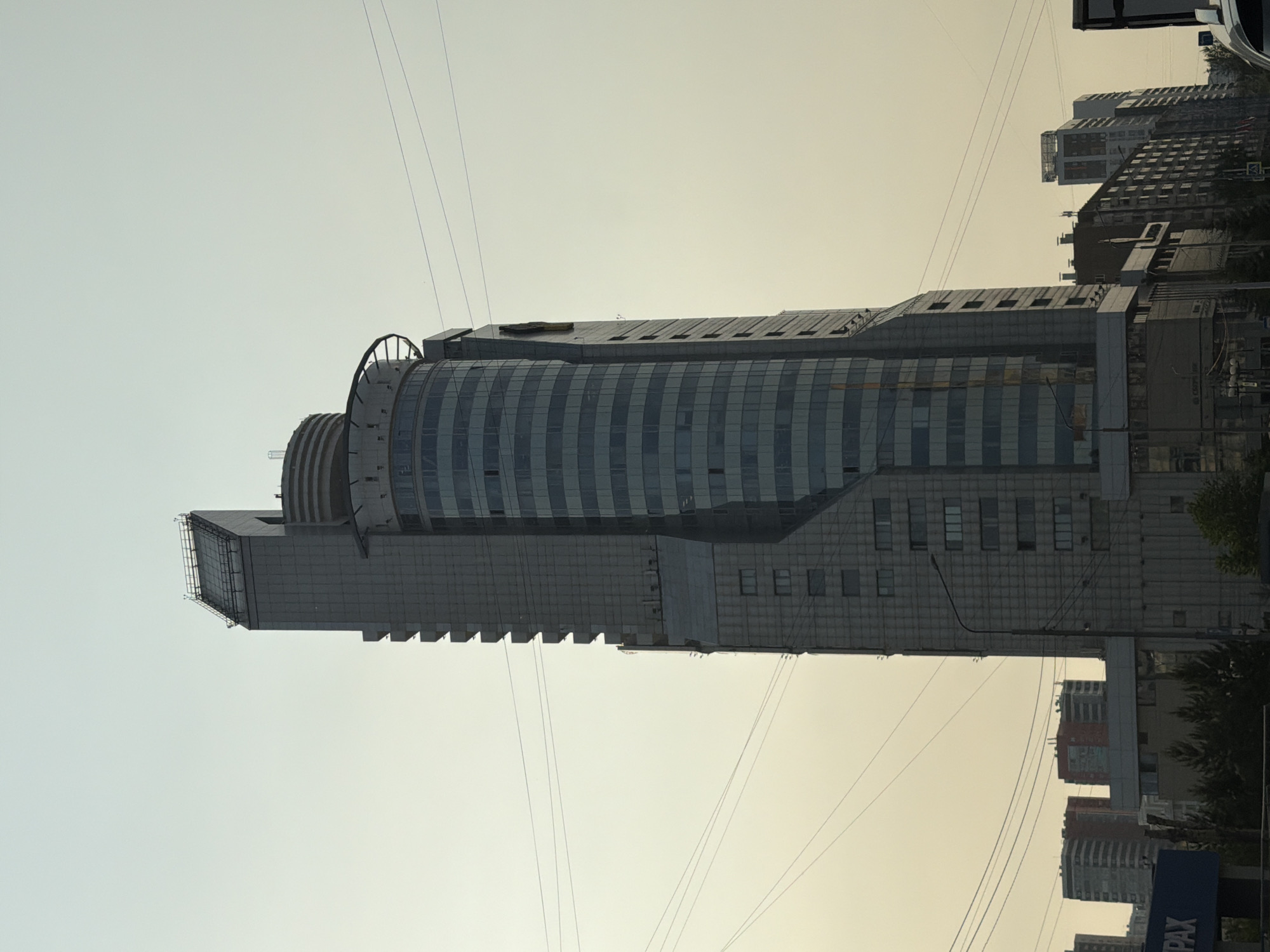 A tall, modern skyscraper dominates the center of the frame, its facade a mix of grey concrete and glass. The skyscraper is angular in its upper sections and curves into a cylindrical glass structure towards its base. Several other buildings of varying heights and architectural styles are visible in the background, some appearing residential and others commercial, typical of an urban cityscape. Overhead, a network of power lines crisscrosses the sky, indicating an established urban infrastructure. The sky is a pale, hazy yellow, suggesting either a sunrise, sunset, or atmospheric pollution. The scene is devoid of any discernible human activity or vehicles, creating a quiet, almost still, urban panorama. The overall impression is one of a bustling city captured during a moment of calm, with the prominent skyscraper serving as a landmark within the urban environment. The image is believed to have been taken in Krasnoyarsk, Russia, specifically near the Vremya Business Center.