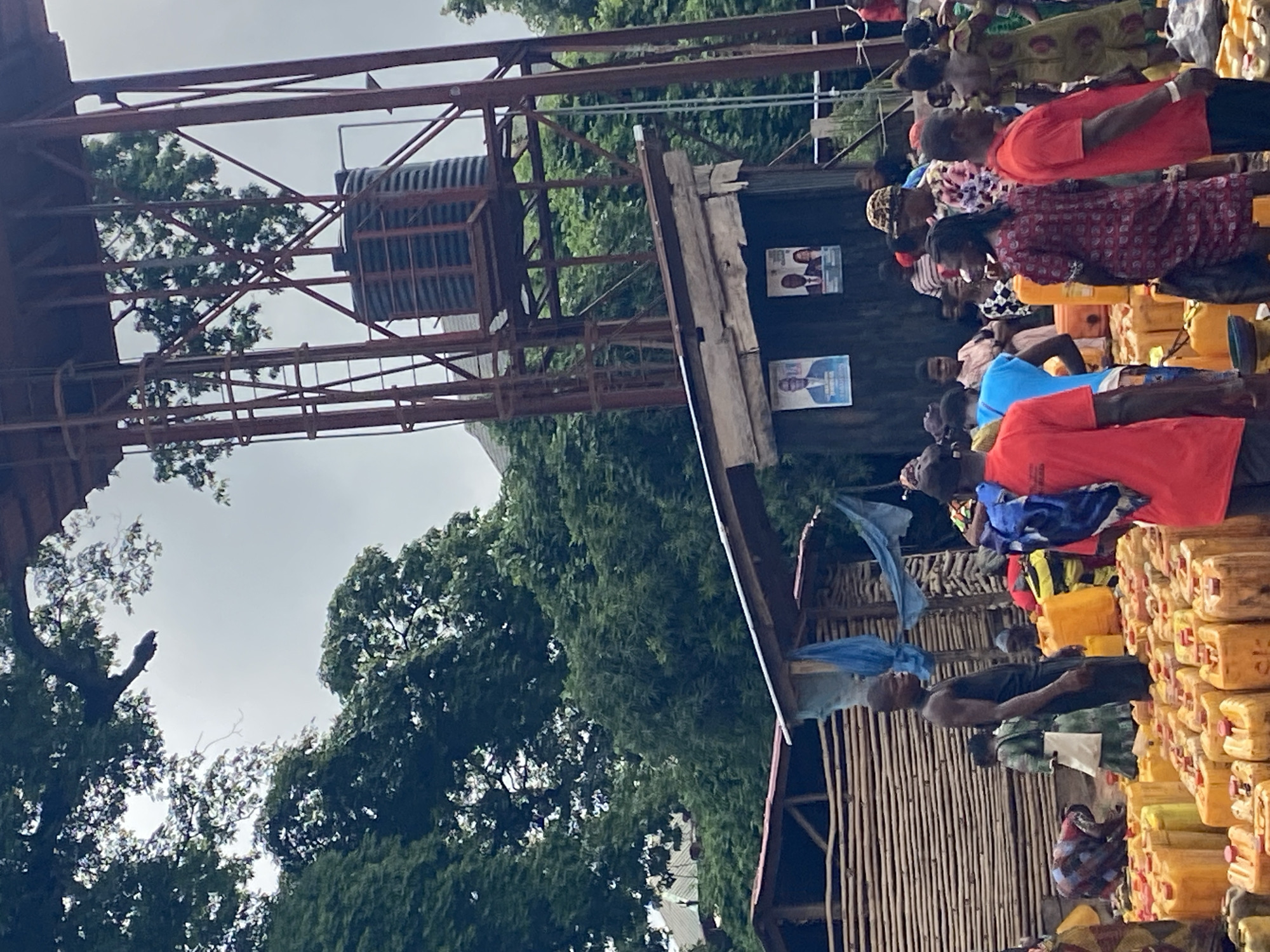 The image depicts a scene in Nguzu Edda, Nigeria, showing a group of people lined up near a water tower or elevated tank structure. The crowd appears to be waiting, possibly to collect water. Many yellow jerrycans are stacked nearby, likely for transporting water. The people are wearing various clothing styles, and some are carrying items. There are political posters on a structure adjacent to the waiting line. The surroundings consist of trees and a building of local architecture, suggesting a rural or semi-urban environment.
