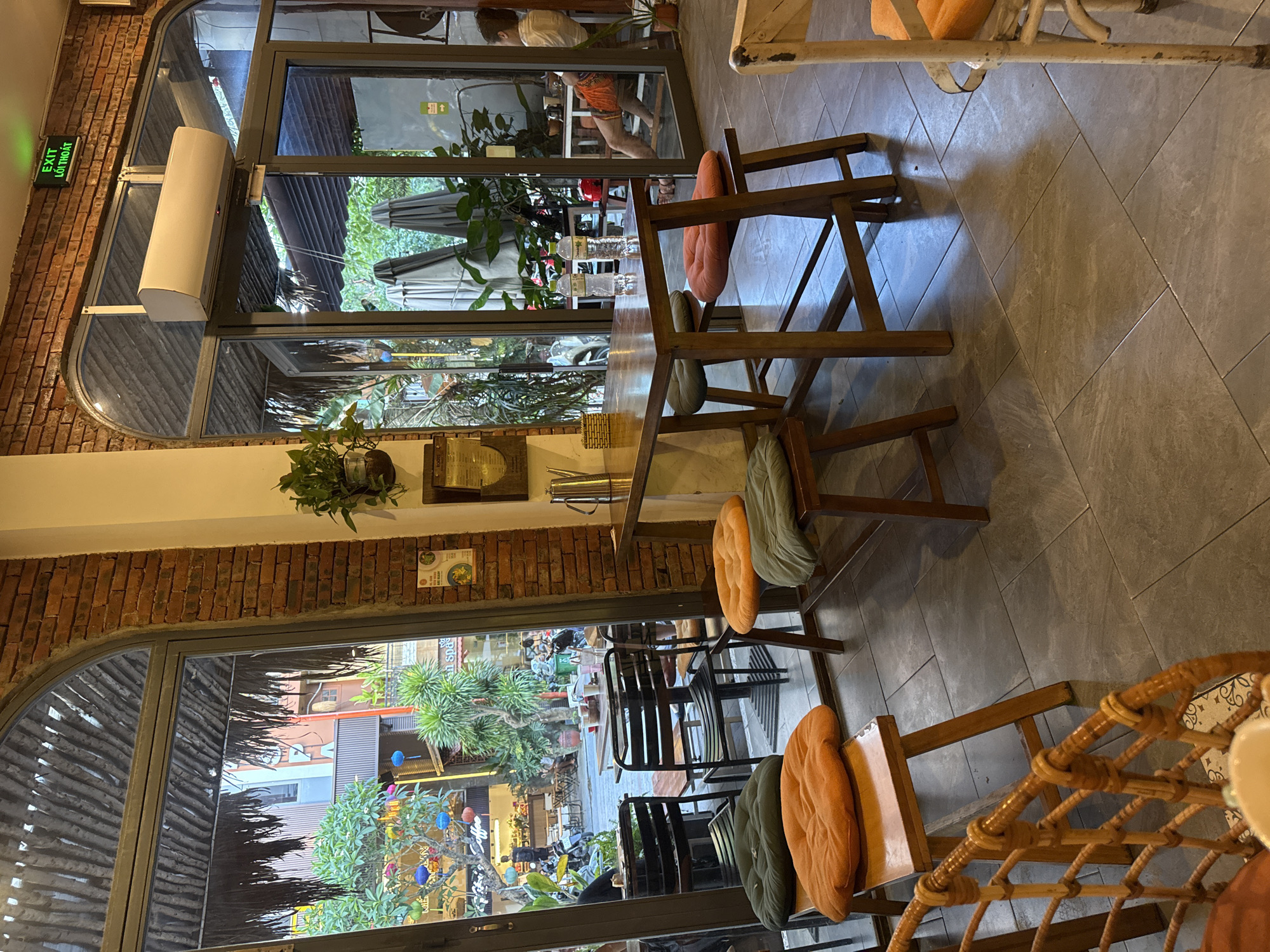 The image shows an indoor cafe setting. The scene is viewed from a slightly elevated perspective, looking out through two large arched windows and glass doors. The cafe features wooden tables and chairs with colorful cushions. Outside, greenery and glimpses of urban life are visible. The overall atmosphere appears relaxed and inviting.