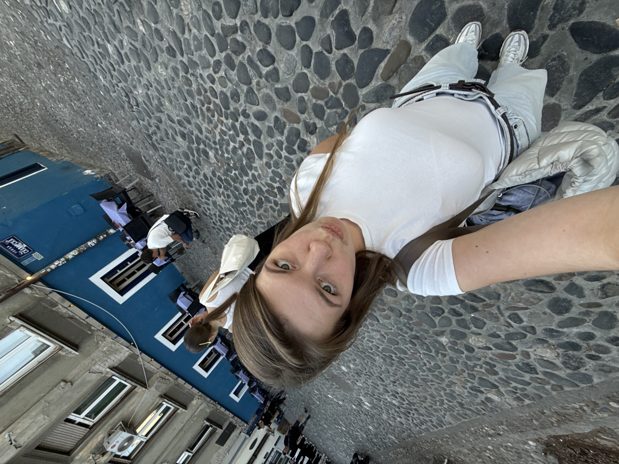 A young woman with long, light brown hair and green eyes is taking a selfie on a cobblestone street. She is wearing a white t-shirt, light blue jeans with a brown belt, and white sneakers. She has a white jacket and gray backpack draped over her left shoulder. In the background, there is a blue building with white window frames, and some people are seated at tables outside. The street appears to be a narrow alleyway in Belgrade, Serbia.