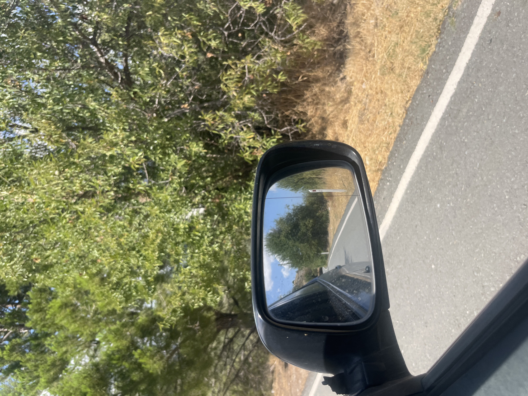 The image is a photograph taken from the passenger side of a vehicle traveling on a paved road. The view is directed towards the side mirror, which reflects a road winding through a landscape of trees and dry grass. The side mirror also shows the sky with some clouds and a white road marker. The vehicle is in motion, as indicated by the blurred road surface and the reflection in the mirror. The environment appears to be a rural or semi-rural area, with lush green trees on one side of the road and dry, yellow grass on the other. The sunlight suggests it is daytime. The location is identified as Silikou, Cyprus. There are no people or specific actions depicted in the image, other than the implied act of driving.