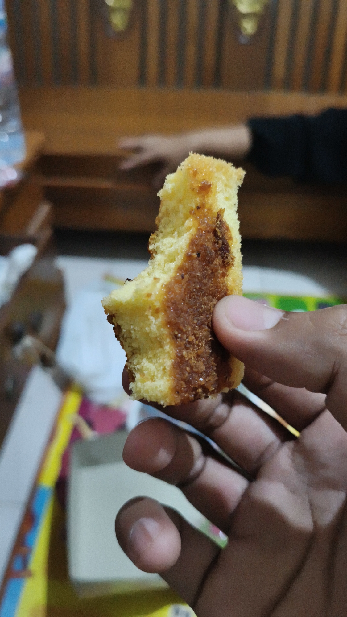 A close-up shot shows a person's hand holding a piece of cake. The cake is light yellow with a darker, caramelized section on the side. The background is blurred, but appears to be an indoor setting with wooden furniture, suggesting a home or restaurant. The presence of a water bottle and packaging hints at a casual, possibly domestic, environment. The lighting is dim and appears to be from an artificial source, casting soft shadows. The image is taken at an angle, focusing on the texture and detail of the cake and the hand.