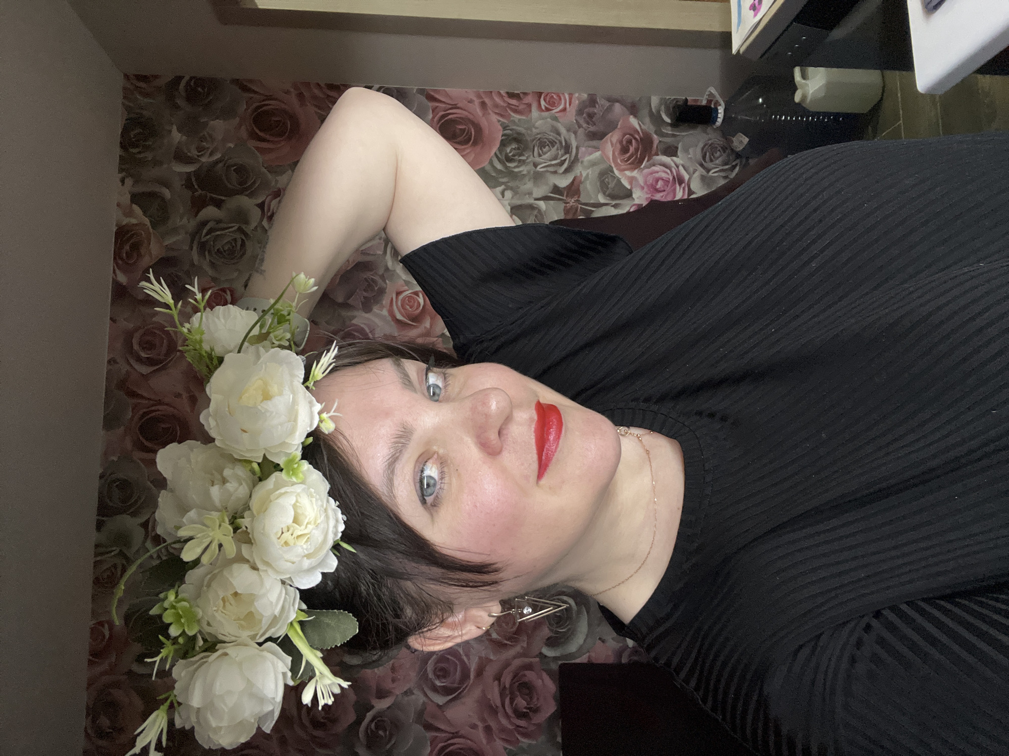 A frontal, slightly angled portrait depicts an adult female indoors in the city of Noviy, Russia. The subject has dark hair, blue eyes, and red lipstick. She is wearing a black, ribbed short-sleeved top, gold-toned triangular earrings with clear stone accents, and a delicate gold-toned necklace. A white artificial floral crown is positioned on her head. Her right arm is raised, with the hand touching the back of her head, near the floral crown. The background consists of wallpaper featuring a repeating pattern of roses in shades of pink, grey, and brown. To the right of the subject, a dark glass carboy with a stopper and a white plastic container are partially visible, situated near a dark surface and a light-colored vertical frame.