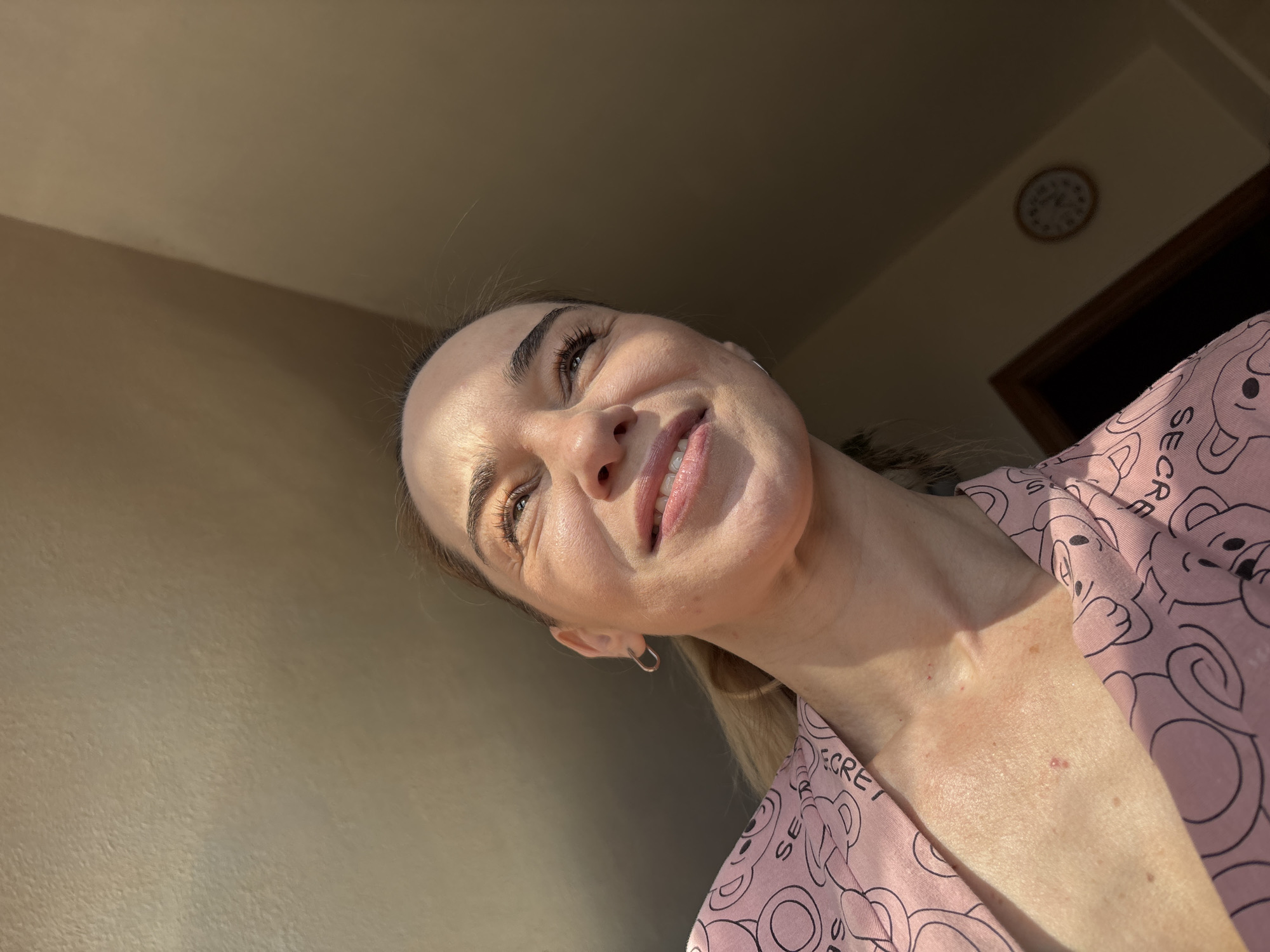 This is a close-up, low-angle portrait of a young woman smiling and looking upwards. She has light skin, dark eyebrows, and her eyes are open and sparkling. Her lips are slightly parted in a smile, and her teeth are visible. She has on a pink robe with a repeating pattern of cartoon teddy bears and the word "SECRET" scattered throughout. She is wearing a small hoop earring in her left ear.
The lighting suggests it is either early morning or late afternoon, as the light is warm and coming from the side, casting shadows and highlighting her features. The background is out of focus but appears to be an indoor setting with a wall and a door. A clock is visible on the wall in the background. The overall mood is cheerful and relaxed.