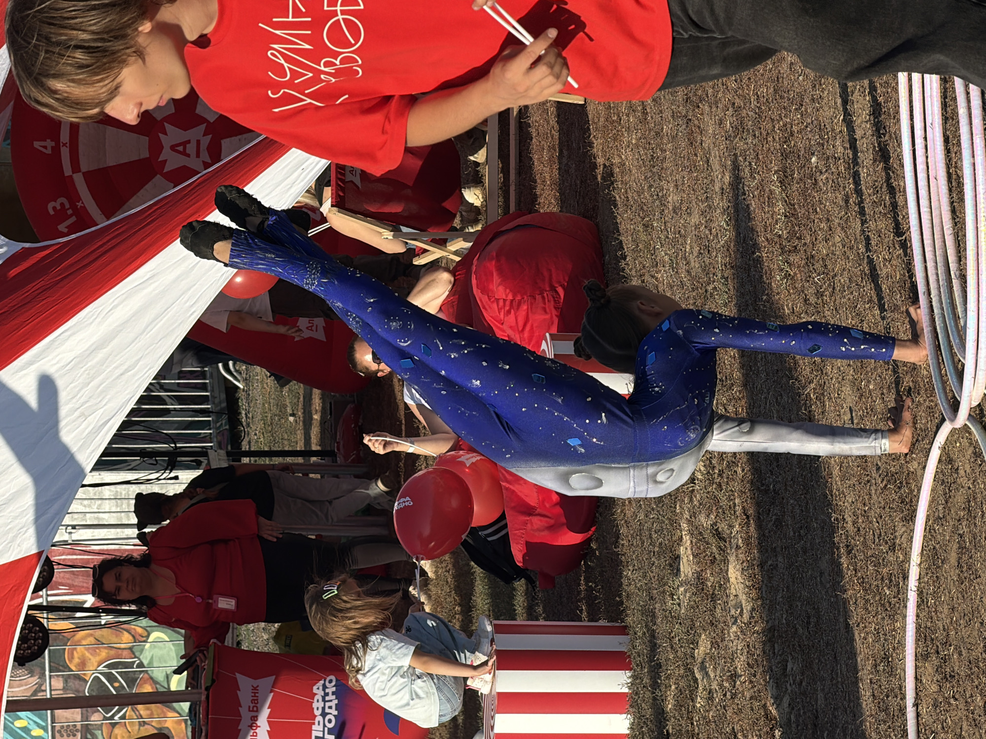 The image captures a street performance or event in Volzhsky, Russia. In the foreground, a young woman in a blue, star-patterned leotard is performing a handstand, with one leg extended upwards. She is positioned on a patch of dry grass, with several hula hoops lying on the ground in front of her.
To the left, a young girl in a light blue t-shirt and jeans crouches on a red and white striped cylindrical platform, holding a red balloon. Behind her, a woman in a red top and dark pants stands near a red stand with Cyrillic text.
In the background, a large red and white tent structure dominates the scene, with some fencing and a large dartboard visible. Other individuals, including a young man in a red t-shirt and dark pants on the right side of the frame, are present, suggesting a public gathering or festival. The overall environment appears to be outdoors, possibly a park or a public square, with dry ground and scattered vegetation. The lighting suggests it is daytime.