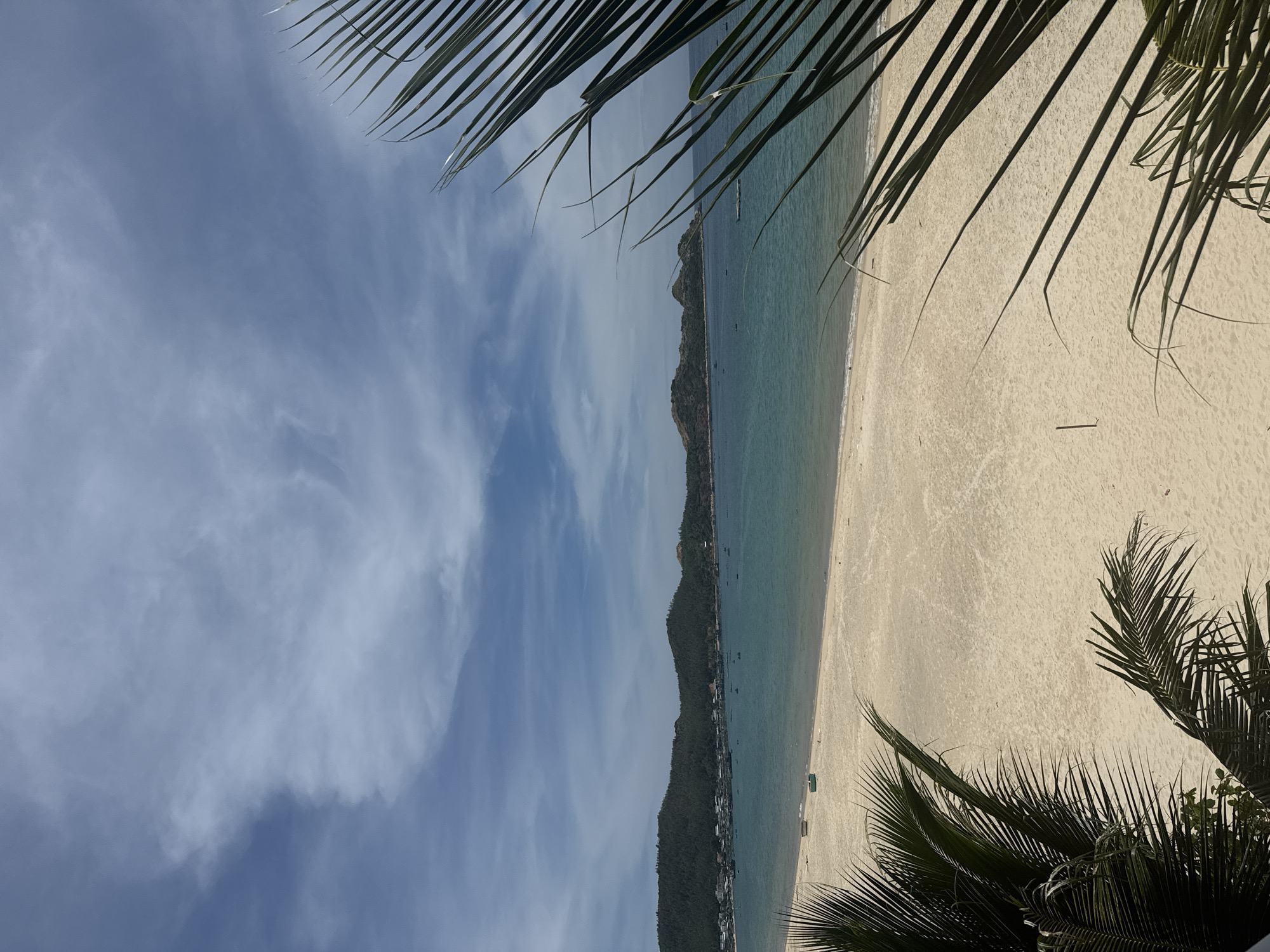 A wide-angle, eye-level outdoor shot shows a tranquil beach scene under a partly cloudy blue sky. In the foreground, the fronds of a palm tree, dark green and silhouetted against the bright sky, frame the view on both the left and right sides. The beach itself is a wide expanse of light-colored sand, marked by subtle ripples and a few scattered shadows from the palm leaves. In the middle ground, the calm, turquoise water meets the shore, with a gentle wave line. Further out, the water deepens to a darker blue. A line of low, verdant, tree-covered hills forms the distant horizon, with a small town or village nestled along the coastline. A scattering of small boats are visible on the water. Two small, light-colored boats are on the sand near the water's edge on the left side.