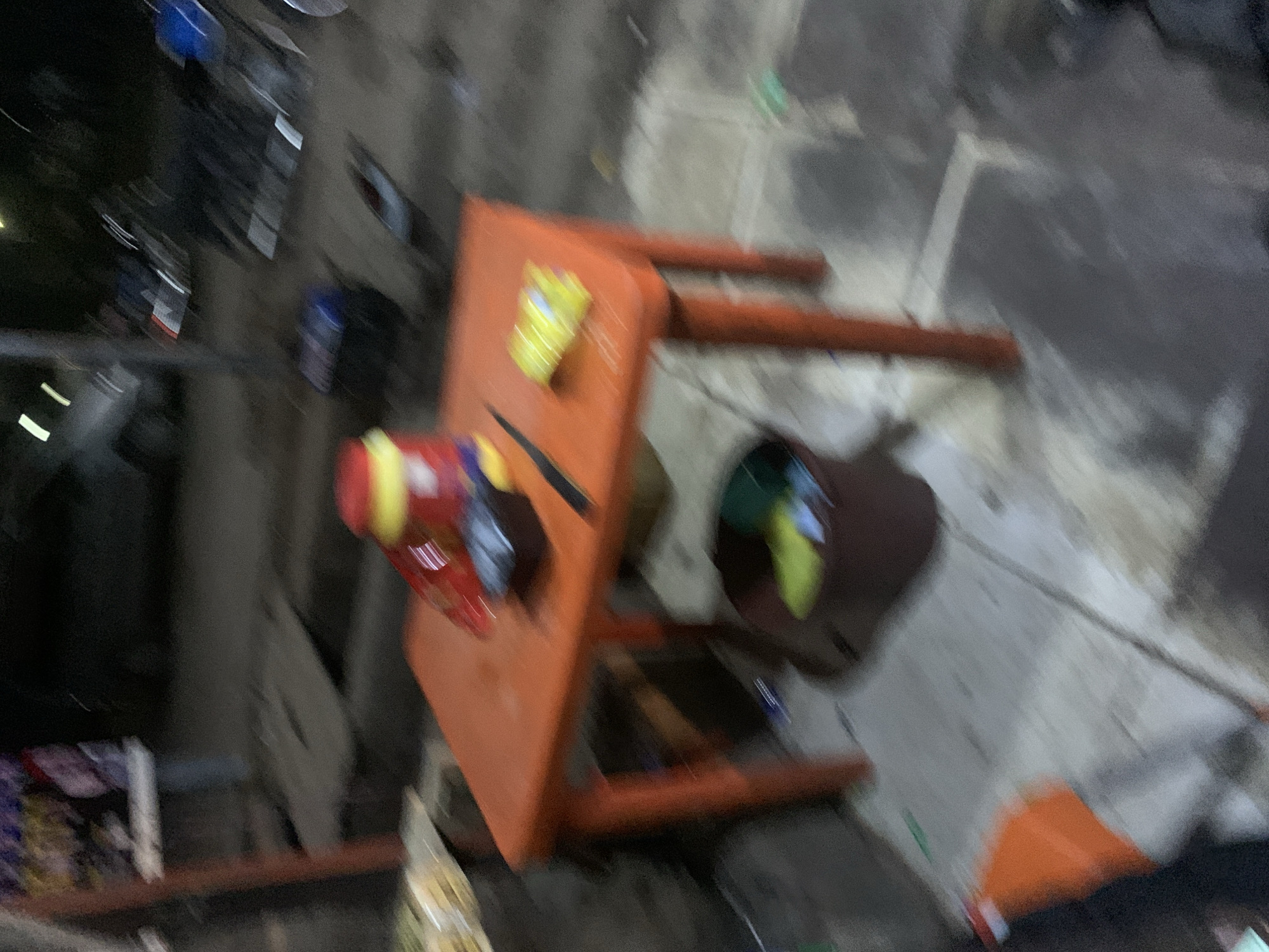 The heavily blurred image depicts an outdoor ground-level scene, possibly an alleyway or an informal market area, viewed from an elevated perspective looking down.
The primary subject is an orange table-like structure situated centrally. On its orange surface, there is a prominent red and yellow object, possibly a container or a toy, and a smaller yellow package. To the right of the table, a dark, cylindrical object, likely a barrel or bin, contains some bright green and yellow items. The ground is a rough, dark grey surface, possibly concrete or pavement, with various indistinct objects scattered around, including a clear bag holding yellowish contents in the bottom-left corner and an orange piece of fabric in the bottom-right. No people are discernible.
The extreme blur across the entire image makes it difficult to identify precise details or specific activities. However, the static arrangement of objects suggests a setup for vending, storage, or a temporary gathering point. The pervasive darkness, combined with what appear to be blurred light sources, strongly indicates that the photo was taken at nighttime or very late evening. No discernible text is visible. The informal setting and arrangement of objects are consistent with scenes found in urban or semi-urban areas.