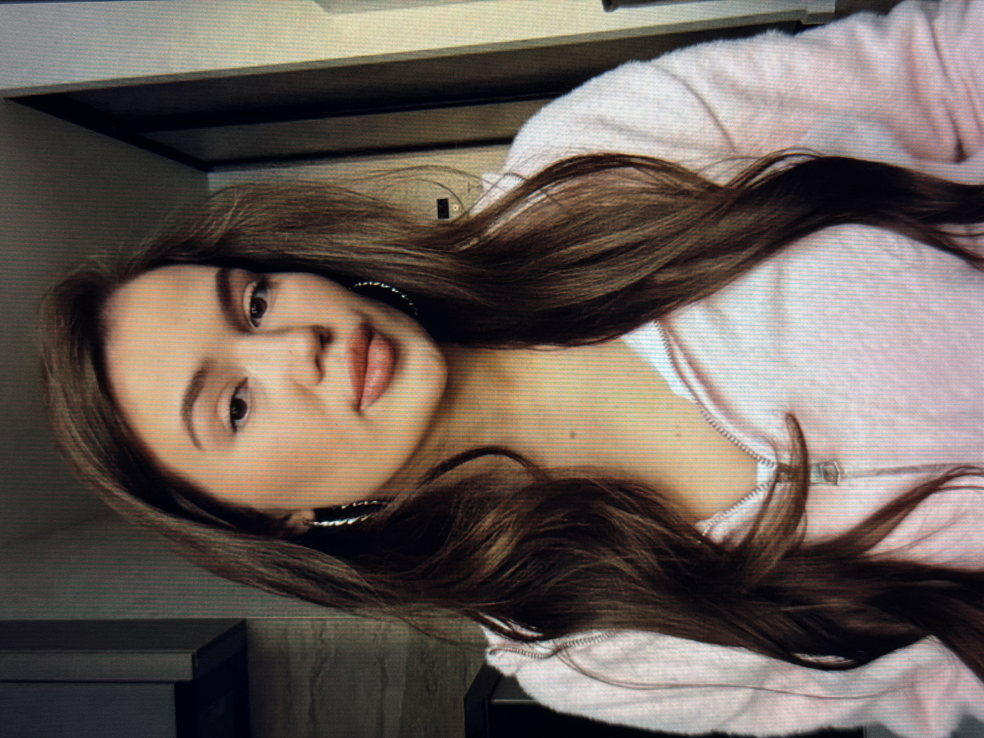 A young woman with long, dark brown, wavy hair is pictured in a close-up, head-and-shoulders shot. She is wearing a light pink zip-up hoodie and large hoop earrings. Her lips are painted a peachy pink, and her eyebrows are well-defined. The background is out of focus, but appears to be an indoor setting with vertical lines suggesting blinds or a door. There is no indication of the city of Sirius, Russia, or any specific location.