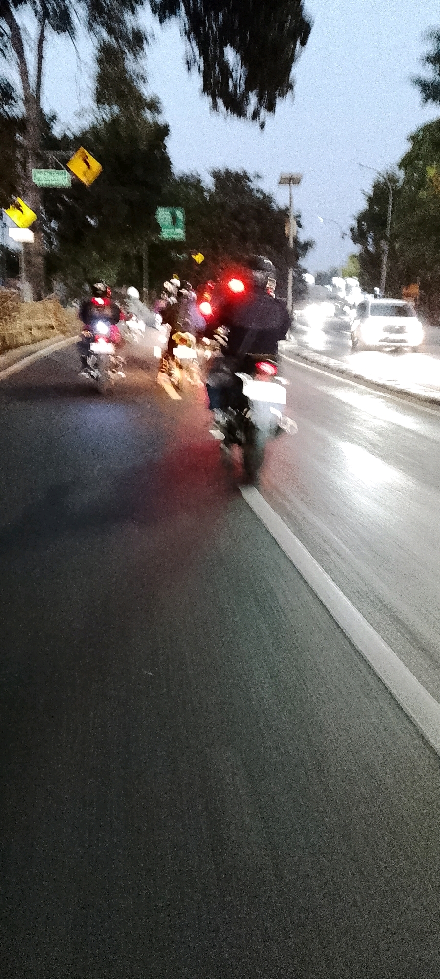 In Depok, Indonesia, a group of motorcyclists are seen riding on a road during twilight or early morning hours. The road appears wet, suggesting recent rain, and the lighting conditions are dim, with the headlights and taillights of the motorcycles and a car in the distance being prominent light sources. The motorcyclists are wearing helmets and jackets, and their bikes have visible taillights. Trees line the side of the road, and several traffic signs are present, including a yellow diamond-shaped sign and a green rectangular sign. The scene is captured with motion blur, indicating movement of the camera or the subjects.