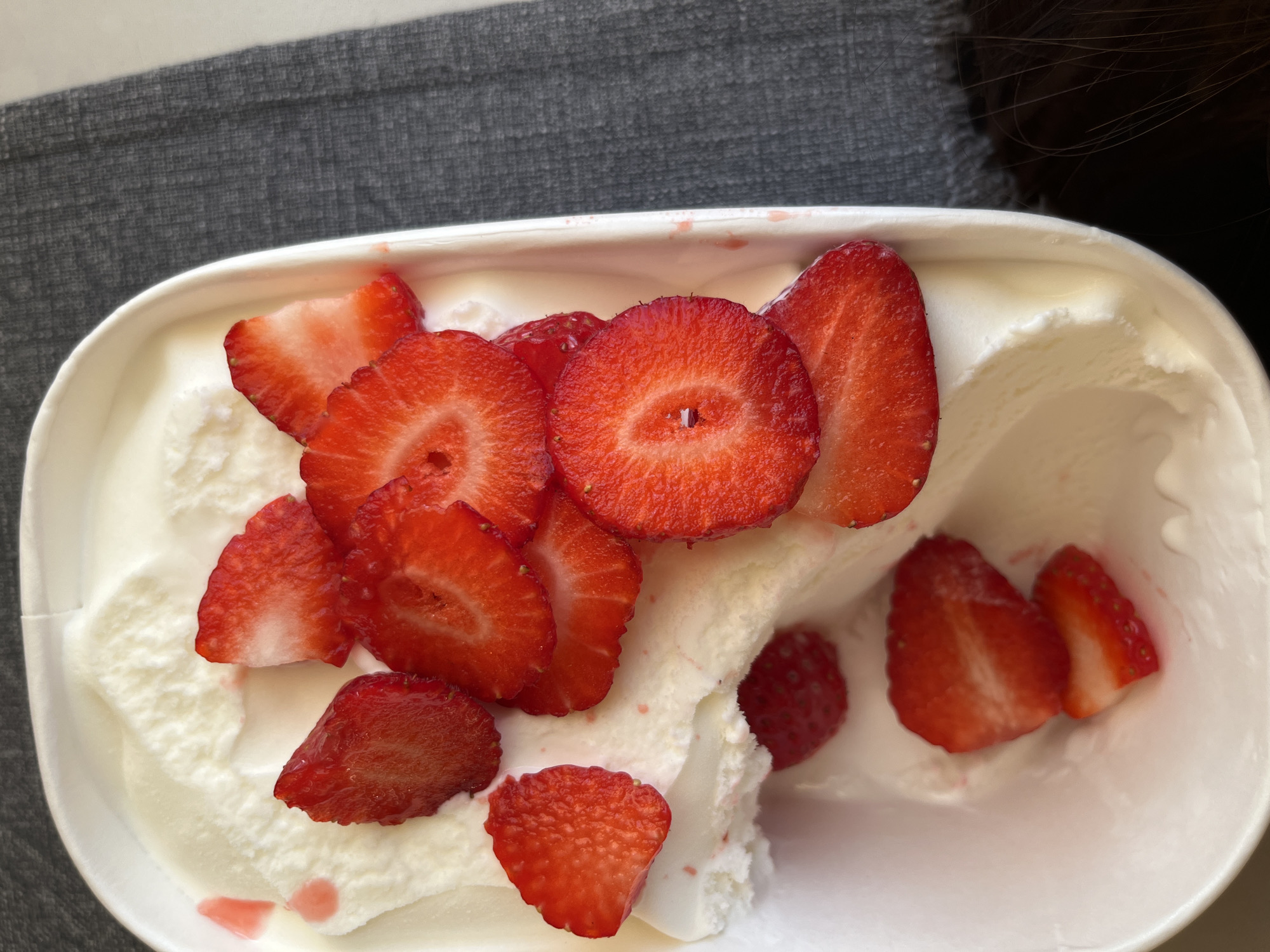 A close-up, eye-level shot displays a white, plastic container filled with white ice cream, topped with sliced strawberries. Some strawberries are cut into rounds, others into wedges. The container sits on a gray, textured surface. A dark, possibly human, hair is visible in the upper right corner of the frame. The setting is assumed to be indoors. The location cannot be determined as Sofia, Bulgaria.