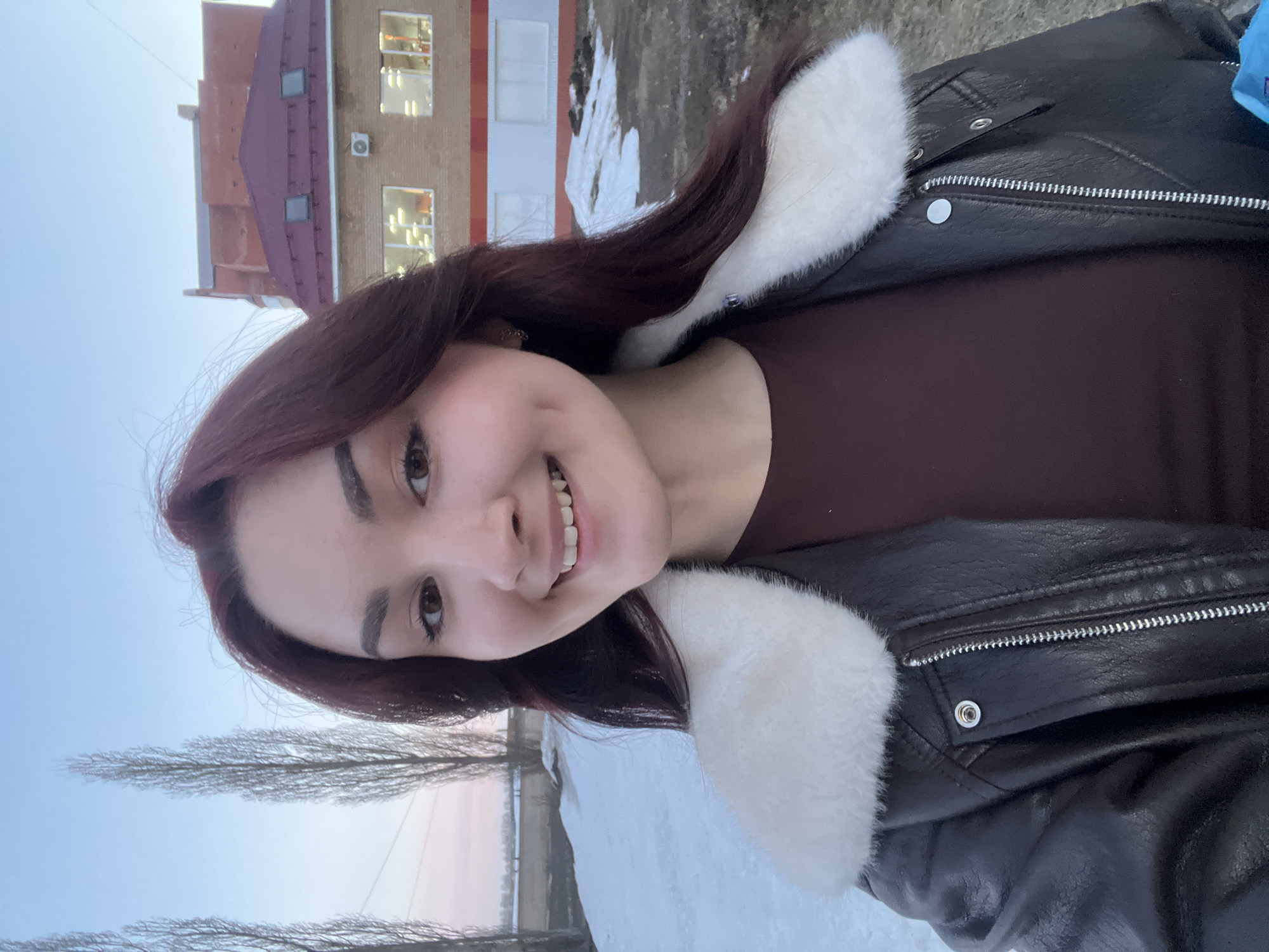 A young woman stands smiling in front of a building in what appears to be a winter landscape in Neftekamsk, Russia. The building behind her is a multi-story structure with a red roof, beige walls, and white trim, possibly an apartment building or office. The ground is covered with snow and bare trees are visible in the background. The sky is pale blue, suggesting either early morning or late afternoon light. The woman is wearing a dark brown, faux leather jacket with a white, fur-like collar and a brown shirt. She has reddish-brown hair and is looking directly at the camera with a smile.