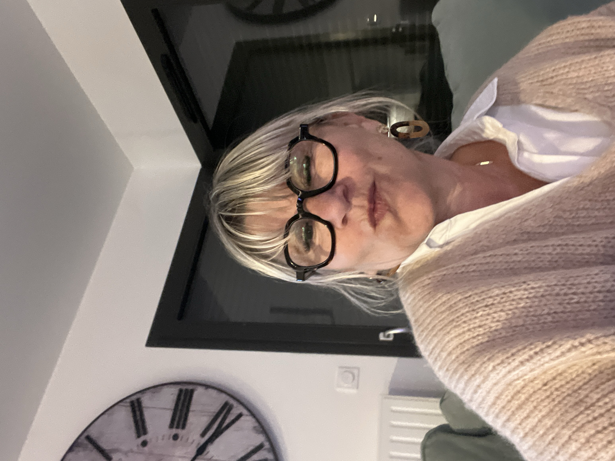 A woman with short blonde hair and black-framed glasses is in the foreground, wearing a white collared shirt and a light brown knitted sweater. Her right ear is visible, sporting a brown and white earring. Behind her is a window reflecting an ambiguous scene. In the background, a large, decorative clock is mounted on a white wall. A small, rectangular electrical outlet is also visible on the wall. The location is Chambœuf, France.