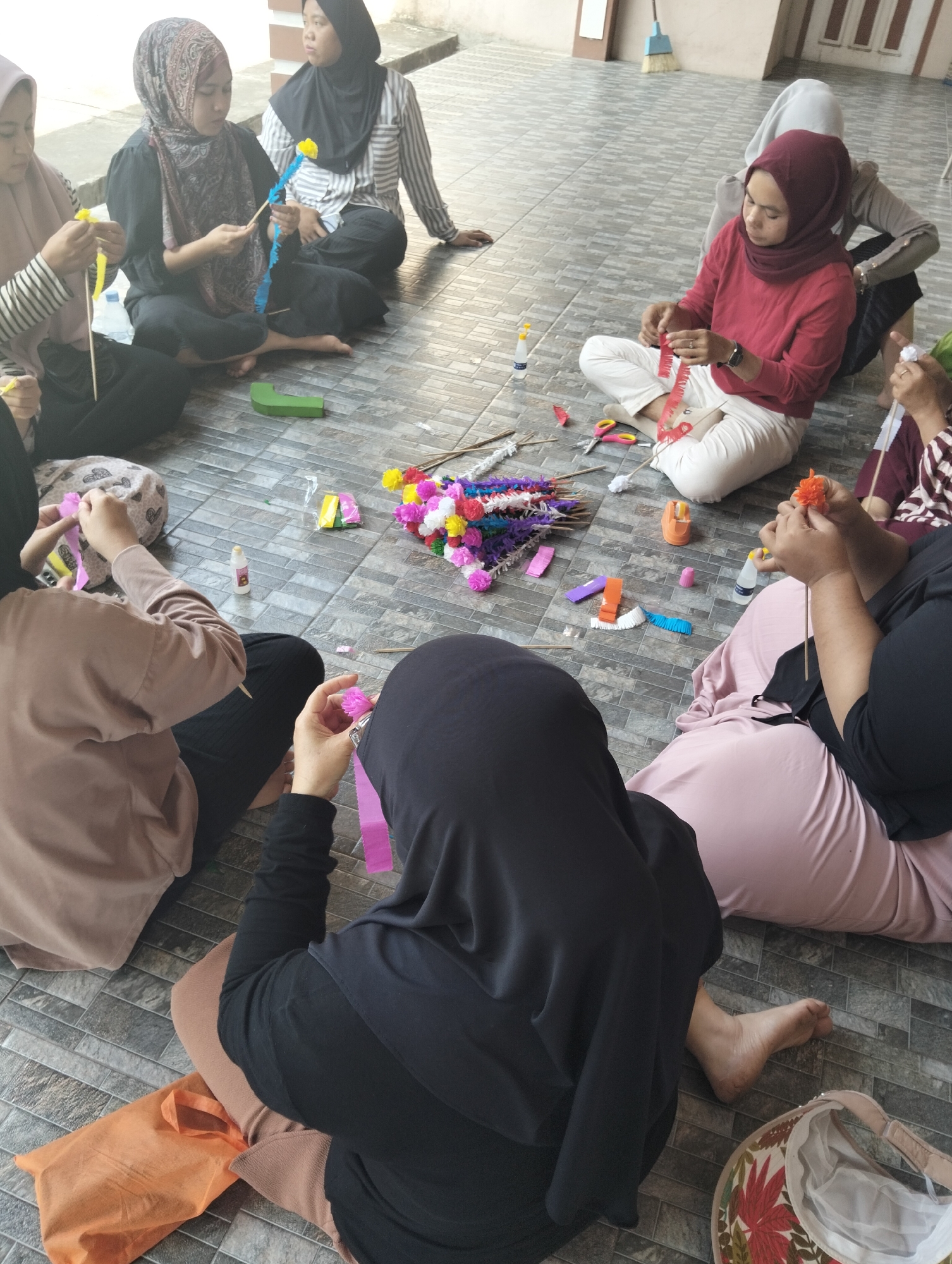 The image shows a group of women sitting on the floor, possibly in Janju, Indonesia, engaged in a craft activity. They are making colorful paper flowers, with various materials like colorful paper strips, scissors, glue bottles, and pre-made flowers scattered around them on the tiled floor. The women are dressed in traditional Islamic attire, including hijabs and loose-fitting clothing. The environment appears to be an outdoor or semi-outdoor space with a tiled floor and a wall in the background. One woman is wearing a maroon hijab and a red top, while another wears a patterned hijab and a black top. Another woman is wearing a light pink hijab and a beige top. A woman in the foreground is wearing a black hijab and a black outfit, actively working on a pink paper flower. The overall scene suggests a community gathering or workshop focused on handicraft.