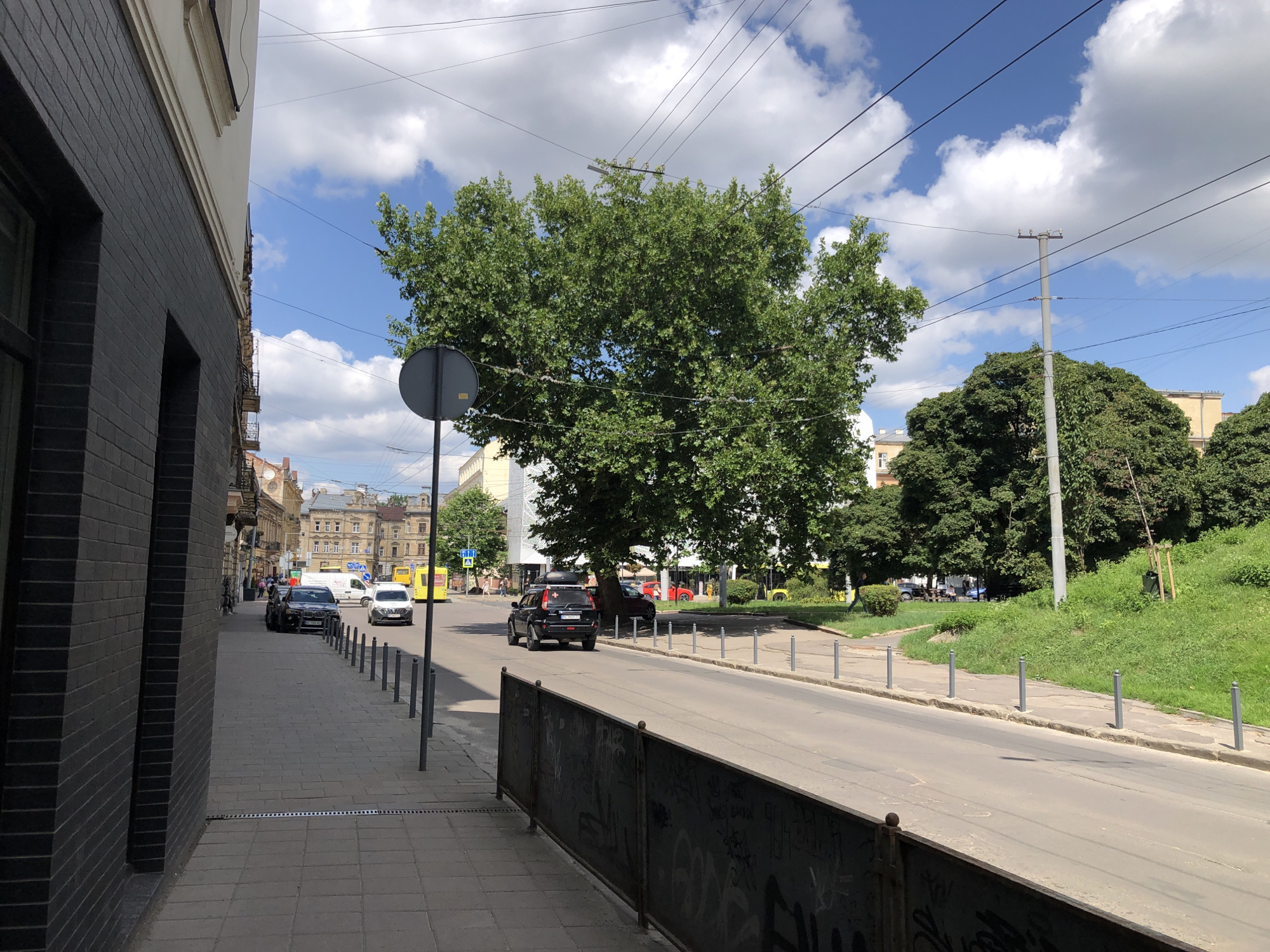 The image depicts a street scene in Lviv, Ukraine, on a sunny day with scattered clouds. A dark brick building is on the left side of the frame, with its wall extending into the foreground. A paved sidewalk runs alongside the building, lined with metal bollards. The street itself is visible, with several cars parked or driving. A large green tree dominates the central part of the image, with its branches reaching across the street. Utility lines are strung across the sky. In the background, there are more buildings, some with ornate architecture, and a grassy slope on the right side of the road. The overall impression is of a typical urban environment with a blend of historical and modern elements.