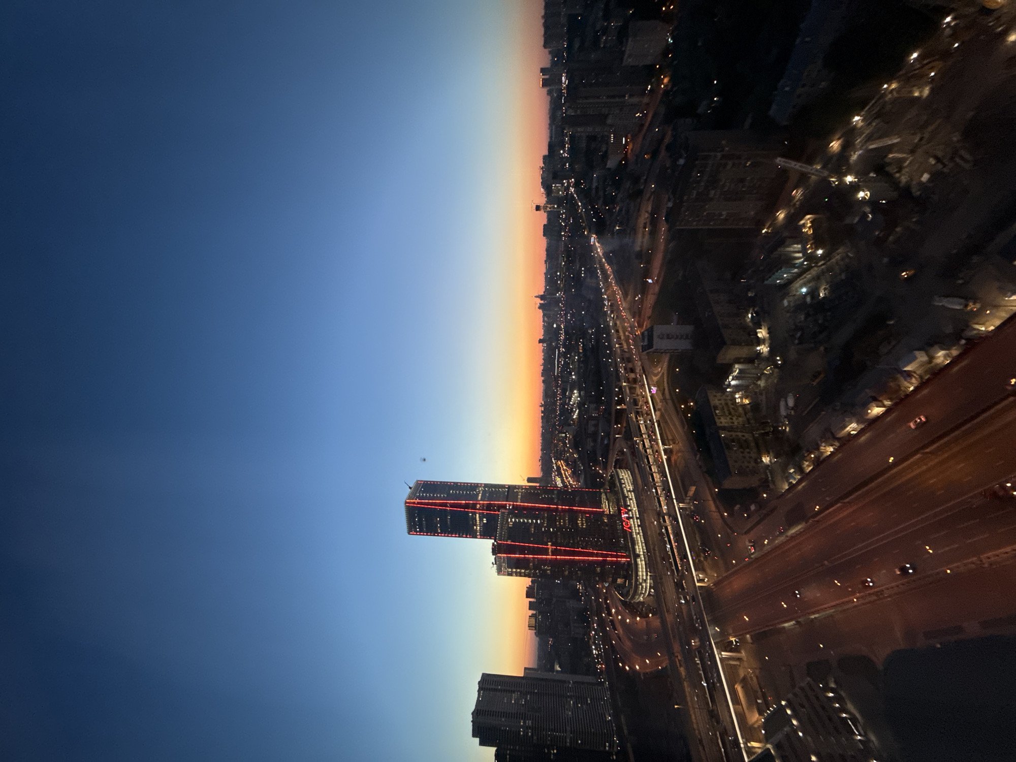 This image captures a panoramic, high-angle view of an urban landscape at twilight, observed from a significant elevation within the city of Khimki, Russia, likely from its southern part near the Moscow Ring Road (MKAD), with the perspective oriented towards the south-southeast. The sky displays a clear gradient from deep indigo at the zenith to vibrant orange and yellow hues along the horizon, characteristic of sunset or sunrise.
In the midground, dominating the central and left portions of the scene, are two modern skyscrapers featuring distinctive vertical red LED lighting elements on their facades. These towers, identifiable as part of the Vegas City Hall / Crocus City complex, rise above a sprawling commercial base, which prominently displays the red "VEGAS" logo. To the immediate left of these central structures, another tall, dark skyscraper is visible, partially silhouetted against the twilight sky.
Below and around these prominent buildings, a complex network of major highways and arterial roads, including segments of the Moscow Ring Road, crisscrosses the urban terrain. These roads are brightly illuminated by numerous streetlights and the headlights and taillights of many vehicles, indicating active traffic flow. Multiple elevated interchanges and road sections are clearly visible, with cars appearing as streaks of light.
The broader cityscape extends to the distant horizon, densely populated with a mix of residential and commercial buildings, all beginning to glow with interior lights and various forms of street illumination. Several construction cranes are discernible in various parts of the distant and mid-ground cityscape, silhouetted or illuminated, suggesting ongoing development. The foreground features additional buildings and segments of the intricate road network, with visible construction sites and work lights in the lower right portion of the frame. The overall scene depicts a vibrant, bustling metropolis transitioning from day to night.