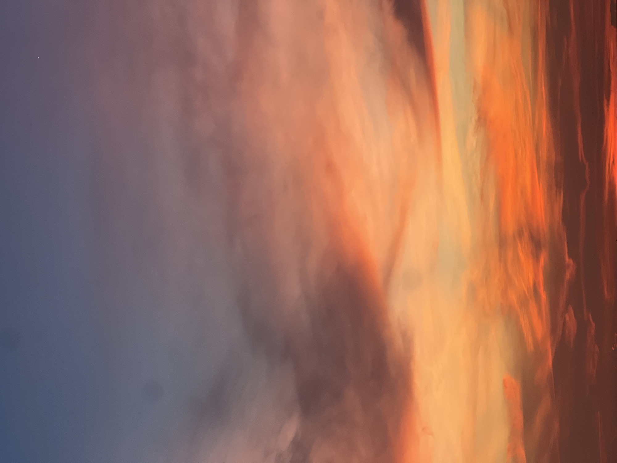 The image depicts a vibrant sunset in Unknown, Unknown. The sky is filled with a mix of colors, ranging from deep blue to vivid orange and red. The clouds are textured and layered, suggesting an active atmosphere. There are no people or recognizable objects visible in the frame, focusing solely on the natural phenomenon.