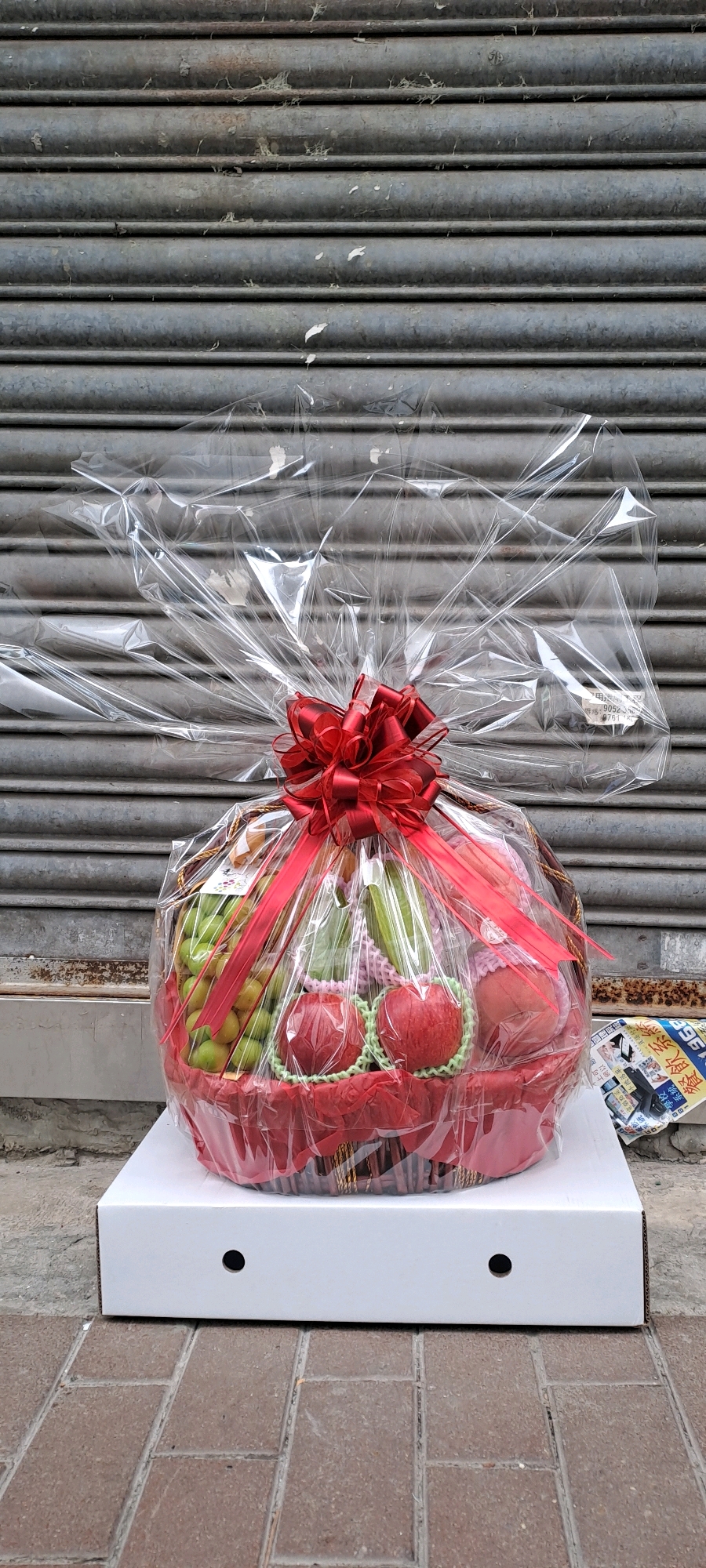 A fruit basket is displayed in front of a closed, grey metal roll-up door. The fruit basket is wrapped in clear cellophane and tied with a red ribbon bow. The basket itself is red and appears to be made of woven material. Inside the basket, various fruits are visible, including green grapes, red apples, and what appear to be green pears or plums. The basket is sitting on top of a white cardboard box. The ground in front of the box is paved with reddish-brown bricks. To the right of the basket, a portion of a newspaper or flyer with Chinese characters is visible. The scene suggests an outdoor setting, possibly a street or alleyway in Hong Kong, China, given the presence of the roll-up door common in commercial establishments and the Chinese text on the flyer. There are no people present in the image. The overall impression is a product display in an urban environment.