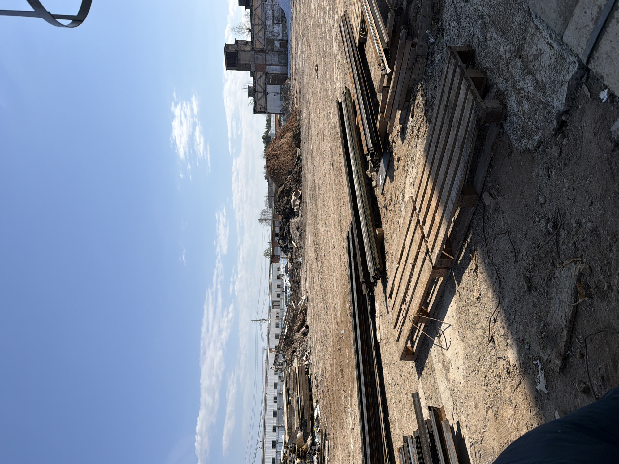 This image captures an expansive view of an outdoor industrial or construction site in Chaikovsky, Russia, bathed in bright daylight under a clear blue sky dotted with scattered white, wispy clouds. The long shadows cast across the ground suggest it is either morning or late afternoon.
The foreground and midground consist of uneven terrain, primarily packed dirt and concrete patches, with piles of loose earth and rubble. Numerous wooden pallets are stacked on the ground, some appearing weathered. Alongside them are many long, dark metal beams or bars, arranged in various piles, indicating stored materials or scrap metal.
In the background, on the left, stand large, decaying industrial buildings. These structures show clear signs of disrepair, with visible brickwork, exposed skeletal frameworks, and missing sections of walls and roofs, suggesting they are either abandoned or undergoing demolition. Further into the distance, a row of long, light-colored buildings, possibly warehouses or factory units, stretches across the horizon, with utility poles and power lines interspersed.
No people are visible in the scene, and no specific activity is taking place. The overall impression is one of an industrial area in a state of transition, disuse, or redevelopment. There is no visible text in the image.