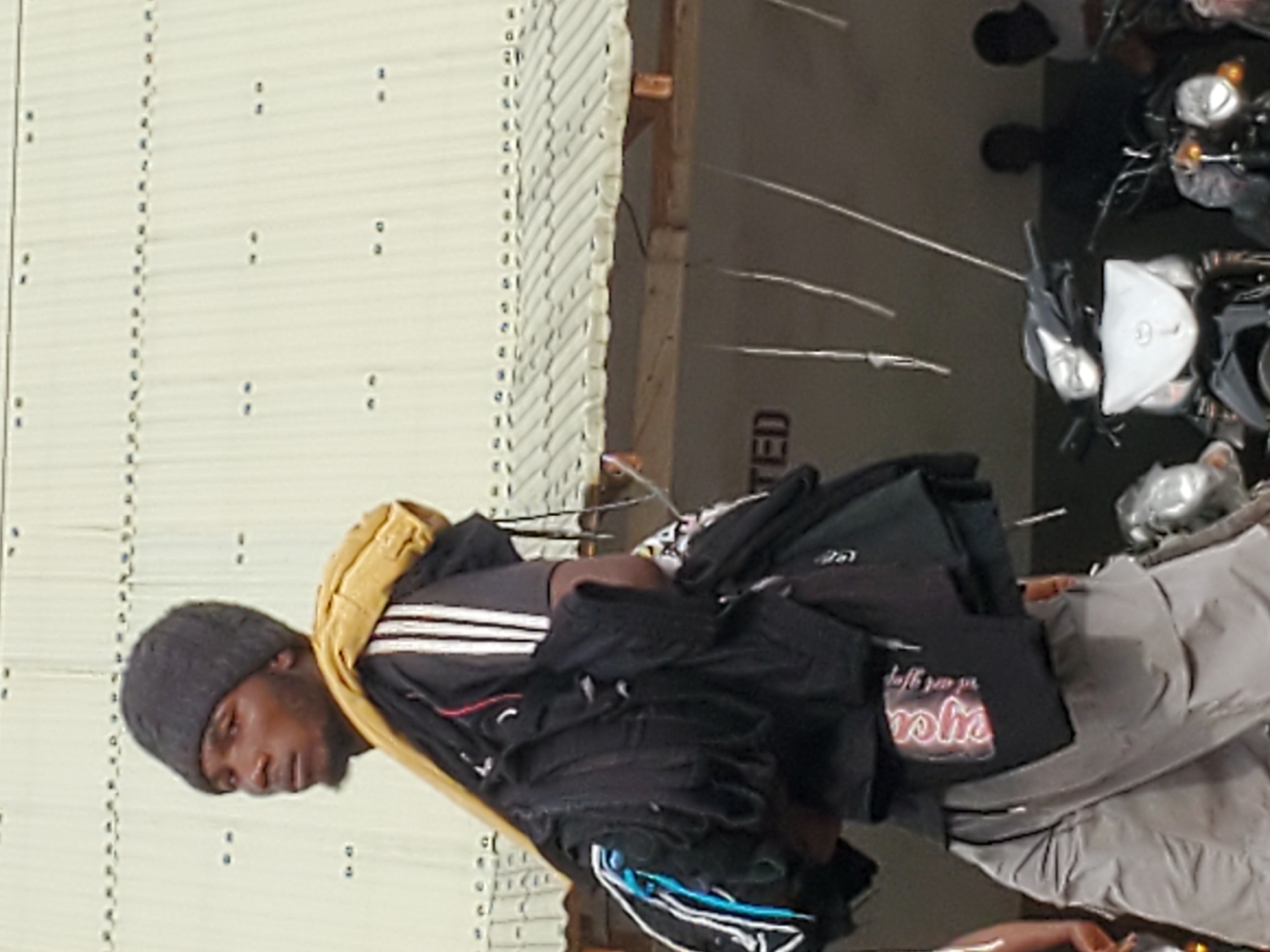 Here is a description of the image.
**People:** An individual wearing a grey beanie, a black Adidas tracksuit with white stripes, a yellow jacket, and grey pants is visible. Their arms are burdened with a large stack of black garments, one with pink writing on it. Two more individuals are seen at the top right, their faces partly visible.
**Objects:** The image captures multiple motorcycles parked in a row. The garments, stacked on the individual's arms, form a notable object. The side of a building is visible, with a series of evenly spaced dots running vertically along its surface.
**Environment:** The setting appears to be an outdoor urban area, possibly a commercial district. The building suggests a commercial establishment. The presence of parked motorcycles implies a street scene. Although the image is rotated, the overall setting appears to be Mararaba, Nigeria.