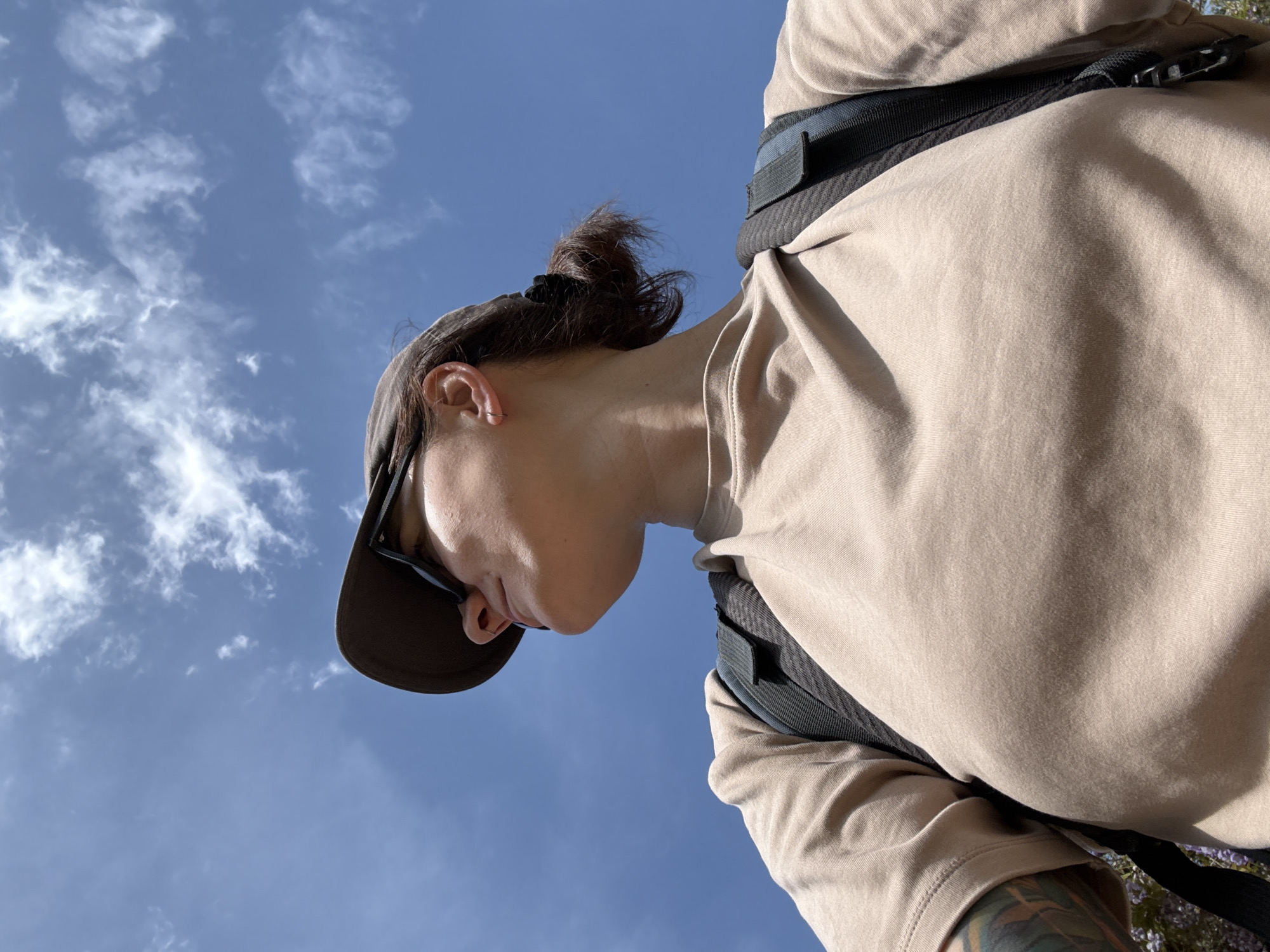 A low-angle shot captures a person wearing a tan shirt, a black backpack strap across their chest, and a dark-colored cap with sunglasses. The person's hair is pulled back into a bun. The background reveals a bright blue sky with scattered clouds. The image appears to have been taken outdoors, possibly in Sveti Stefan, Montenegro.