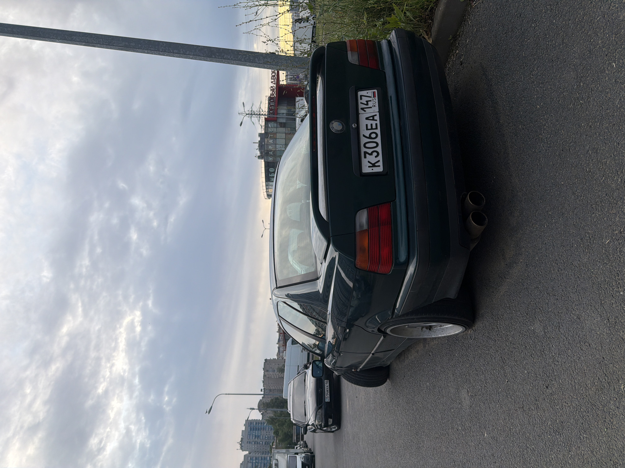 A dark green sedan is parked on an asphalt road in an urban environment. The car is angled towards the viewer's left, with its rear facing the camera. The license plate reads "K306EA 147" and has a Russian flag symbol. The car has a spoiler on the trunk and dual exhaust pipes. In the background, there are buildings, streetlights, and other vehicles, including a black sedan and white vans. The sky is overcast with clouds. The scene appears to be a street or parking area in a city.