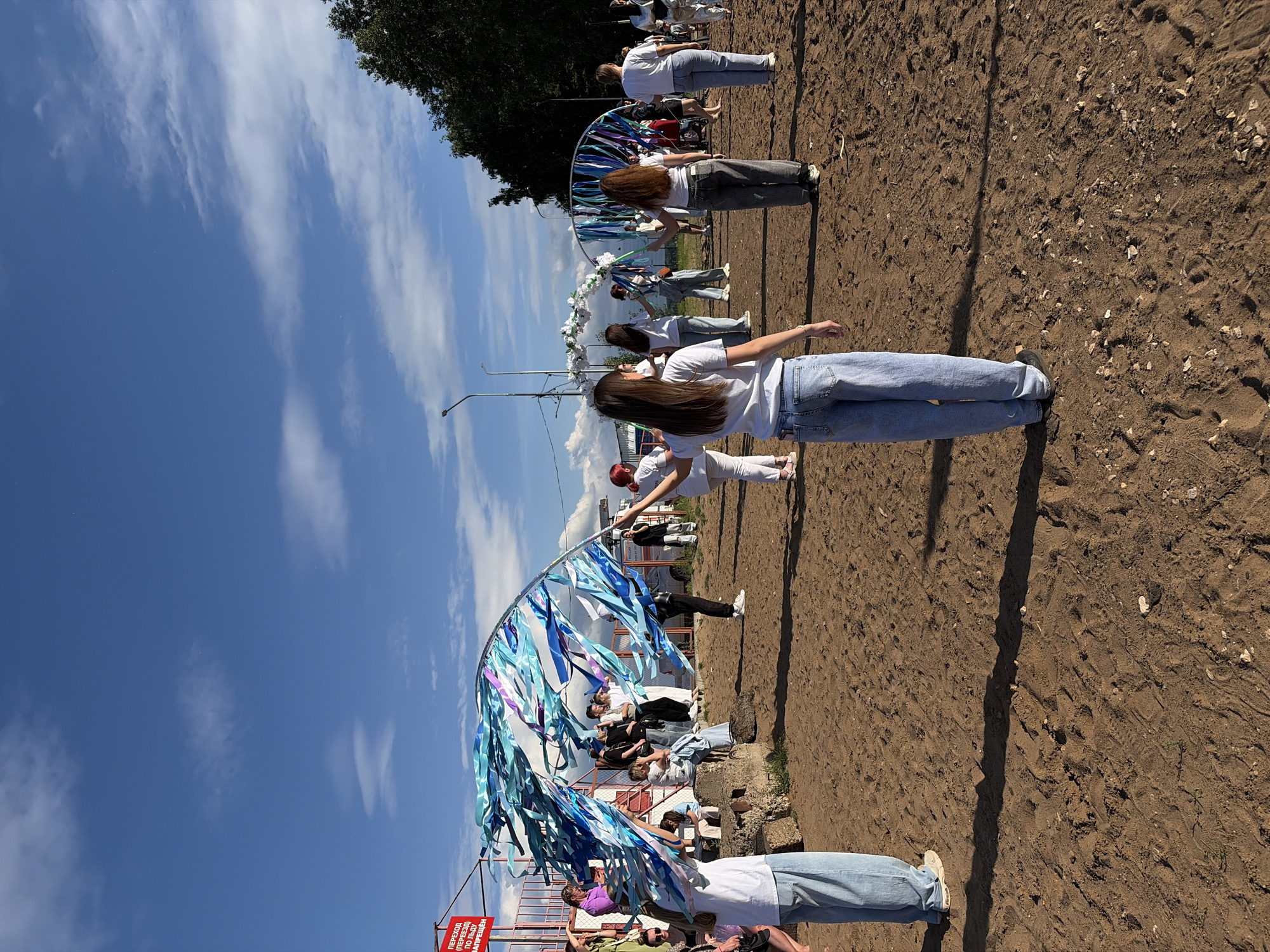 In Chaikovsky, Russia, on a bright, cloudy day, a group of young people are engaged in what appears to be an outdoor performance or rehearsal on a sandy area. Several individuals, mostly dressed in casual attire including t-shirts and jeans, are holding and manipulating large, decorative arcs adorned with blue and white streamers, possibly for a dance or festival. One person in the foreground, facing away from the camera, is holding a streamer-covered arc. In the background, spectators are seated on a concrete structure, and a red sign with Russian text is visible on a metal fence. The environment suggests a public space, perhaps a park or a designated event area, with trees and a street lamp in the distance.