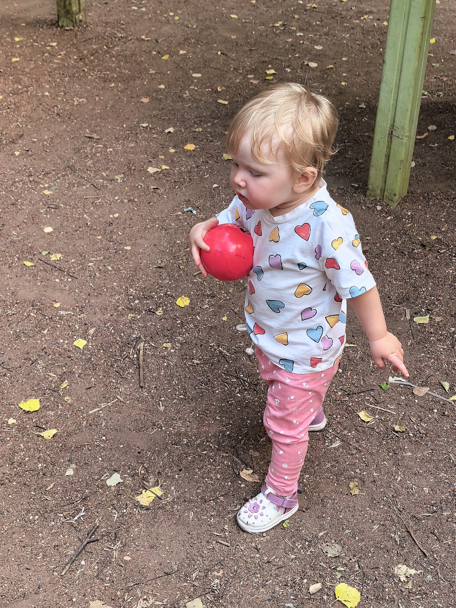 A young child with blonde hair is standing on a dirt surface with scattered leaves and small stones. The child is wearing a white t-shirt with colorful heart patterns, pink leggings with a floral print, and white and purple sandals. The child is holding a red ball in their right hand and pointing with their left index finger towards the ground. In the background, a green, weathered post is visible. The scene appears to be outdoors in a park or playground area. The image was captured in Sterlitamak, Russia.