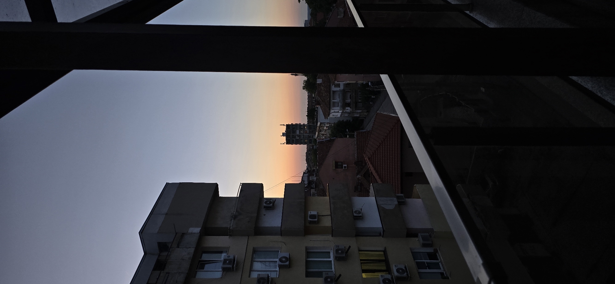 The image depicts an urban environment at dusk from an elevated perspective, likely a balcony. The sky exhibits a gradient from muted blue-grey directly overhead to vibrant orange and pink hues at the horizon. In the immediate foreground on the right, dark structural elements and a silver-colored balcony railing are visible, partially obscuring the view. To the left, a multi-story residential building with a light yellow exterior features multiple balconies and numerous external air conditioning units; one lower-level window on this building emits a warm yellow light. Further into the midground, a dense arrangement of buildings with traditional red-tiled roofs and a visible street are present. A tall, dark, multi-story building, appearing as a silhouette with antenna-like structures on its roof, stands prominently against the sunset on the distant horizon. No human figures are discernible within the scene. The overall lighting suggests the late evening transition from daylight to night.
