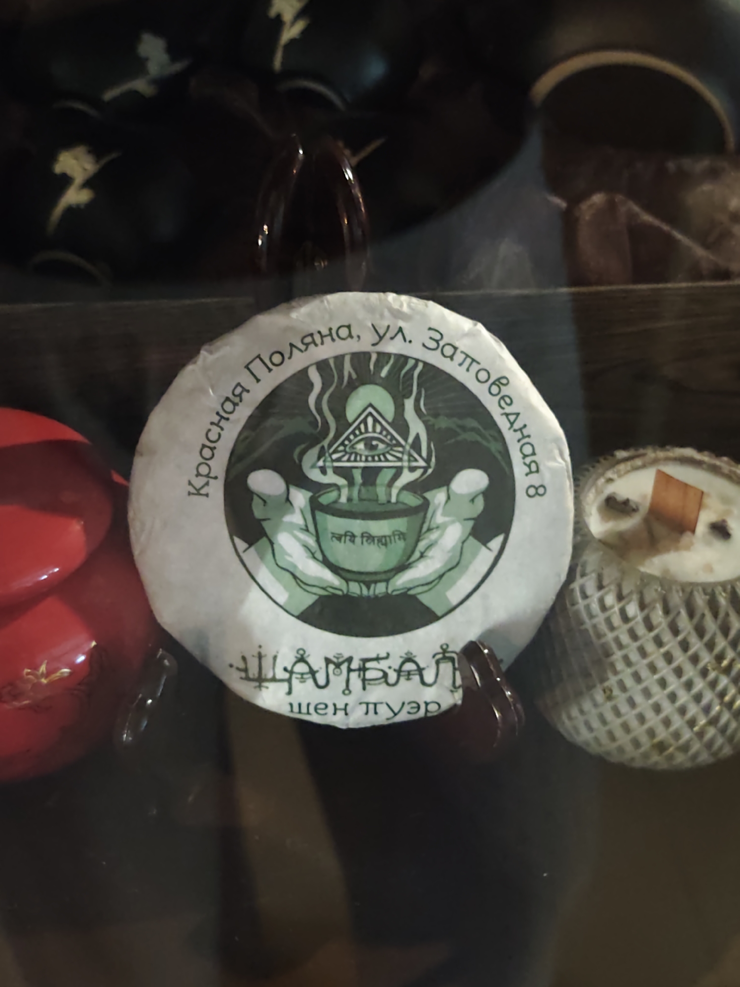 The image shows a close-up of a round, white tea cake with a green design in the center. The design depicts a stylized eye within a triangle, above two cupped hands holding a bowl that is emitting steam. There is text in Russian around the circular border of the design, reading "Красная Поляна, ул. Заповедная 8". Below the design, in Cyrillic script, are the words "ШАМБАЛ" and "шен пуэр". To the right of the tea cake is a cylindrical object wrapped in a woven material, with a wooden wick and what appears to be dried herbs or spices visible within a creamy substance. In the background, dark, possibly lacquered objects with decorative gold patterns are visible. A red, glossy object is partially visible on the left side of the frame. The overall scene appears to be a retail display, possibly in a specialty tea shop or market. The text "Красная Поляна, ул. Заповедная 8" indicates the location as the address "Zapovednaya Street 8, Krasnaya Polyana". Krasnaya Polyana is a mountain resort town located in the Sochi municipality, Krasnodar Krai, Russia, known for its ski resorts.