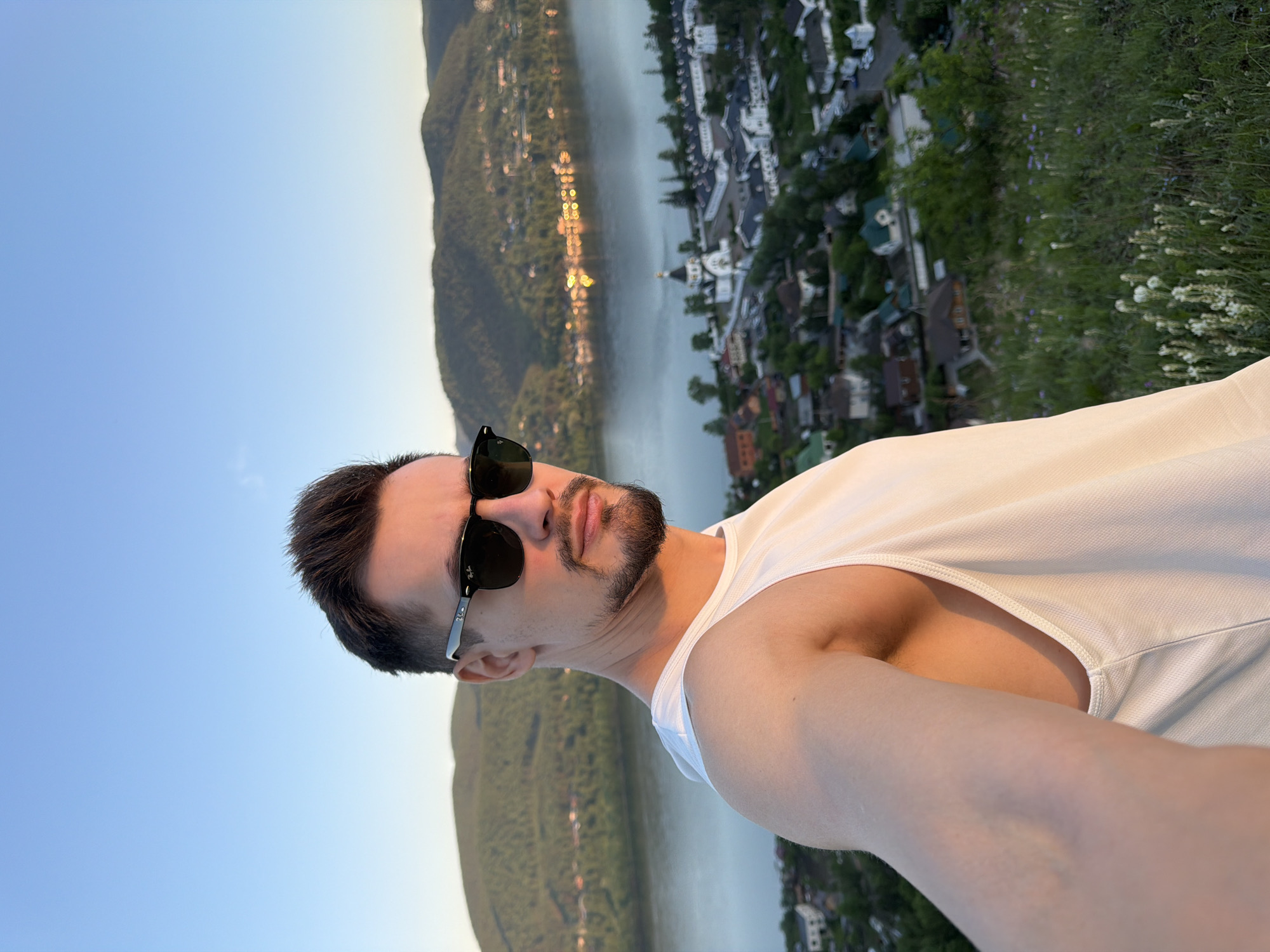 A man is depicted in a selfie, taken from a high vantage point overlooking a city, river, and mountainous landscape during the golden hour. The man, with dark hair styled upwards, a dark beard, and mustache, wears black Ray-Ban sunglasses and a plain white sleeveless tank top. He faces the camera with a neutral expression, positioned slightly to the left of the frame's center. His right arm, holding the camera, extends into the lower left corner. Golden sunlight illuminates the right side of his face and shoulder.
The background reveals a panoramic view. In the immediate foreground, a grassy hillside with sparse small white and purple wildflowers descends. Below, the Yenisei River flows, reflecting the blue sky. On the river's far bank, a prominent white monastic complex with multiple black-domed churches and associated buildings (the Holy Dormition Monastery) is clearly visible, surrounded by residential structures and green trees. Further beyond the river, densely forested hills and mountains rise under a clear, light blue sky transitioning to a warmer hue near the horizon. Lights from a settlement are reflected on the river's surface in the middle-right. The scene is located in Krasnoyarsk, Russia, with the viewpoint likely from a hill offering an overview of the Yenisei River and the Holy Dormition Monastery.